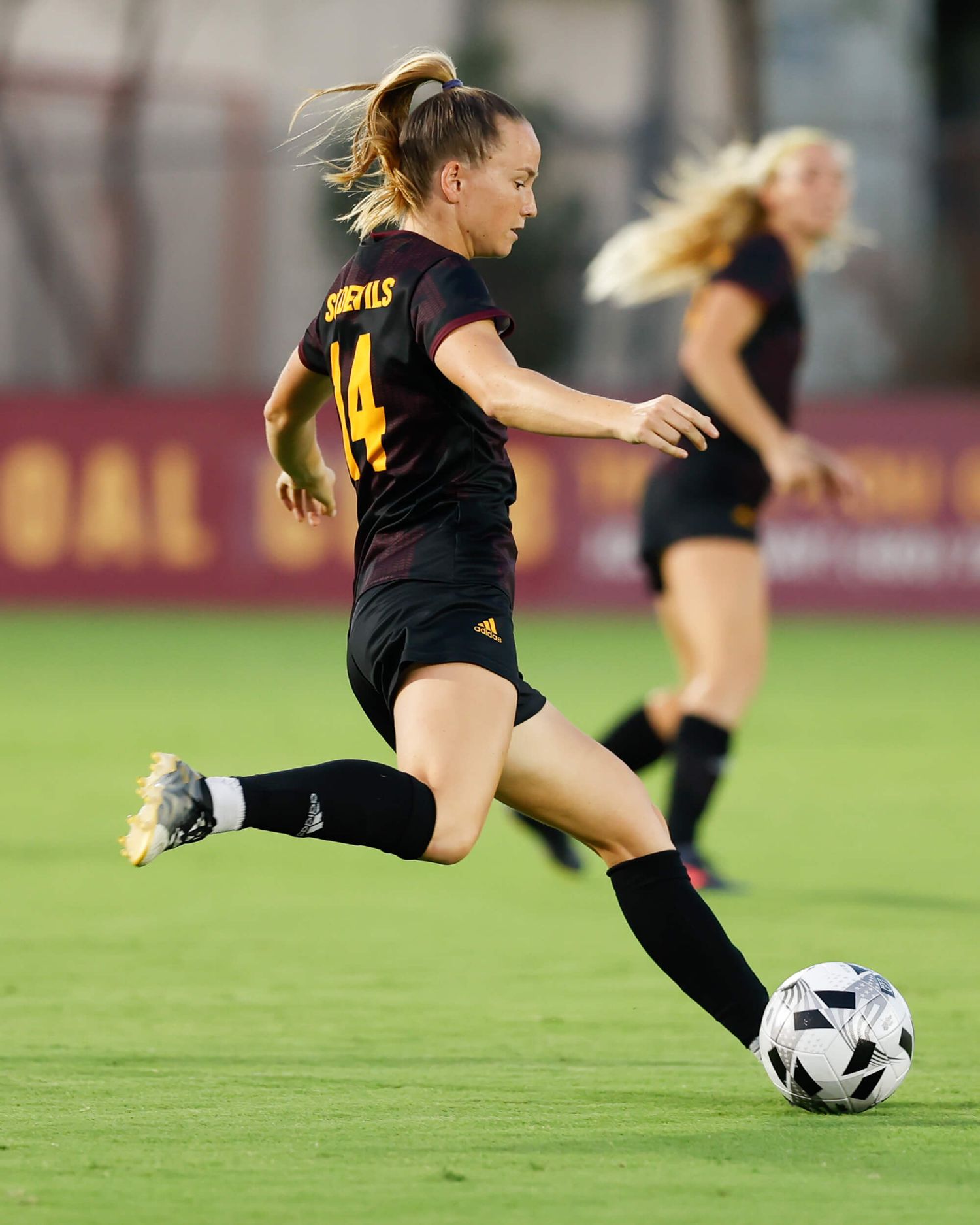 Female soccer player in maroon uniform number 14 prepares to kick ball during evening college soccer match.