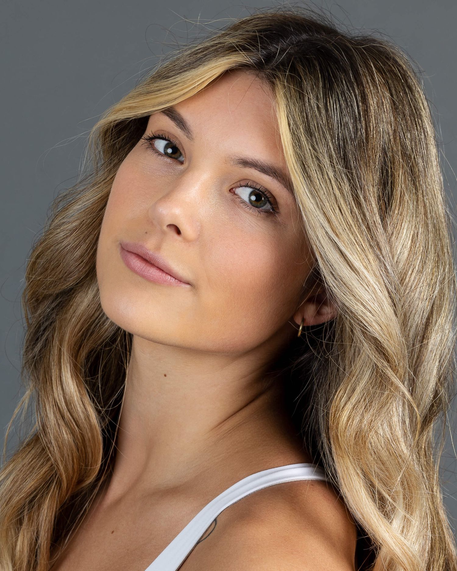 Young blonde woman with wavy hair wearing a white top poses for a professional modeling headshot.