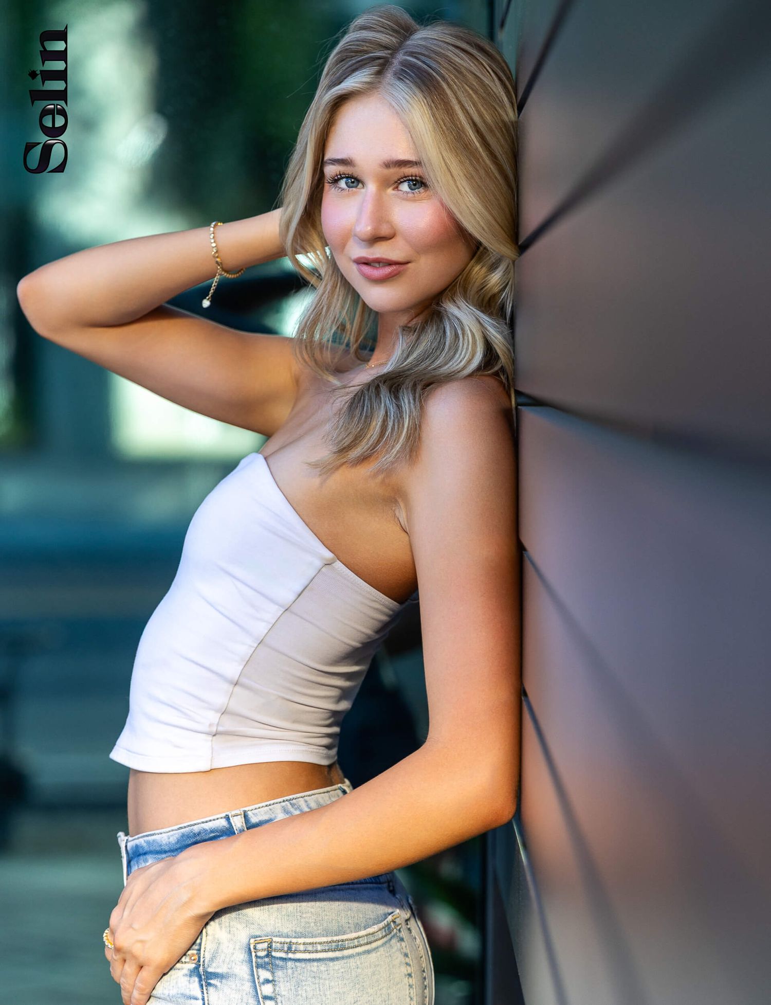 Blonde model in white tube top and jeans leans against dark wall for Selin magazine shoot.