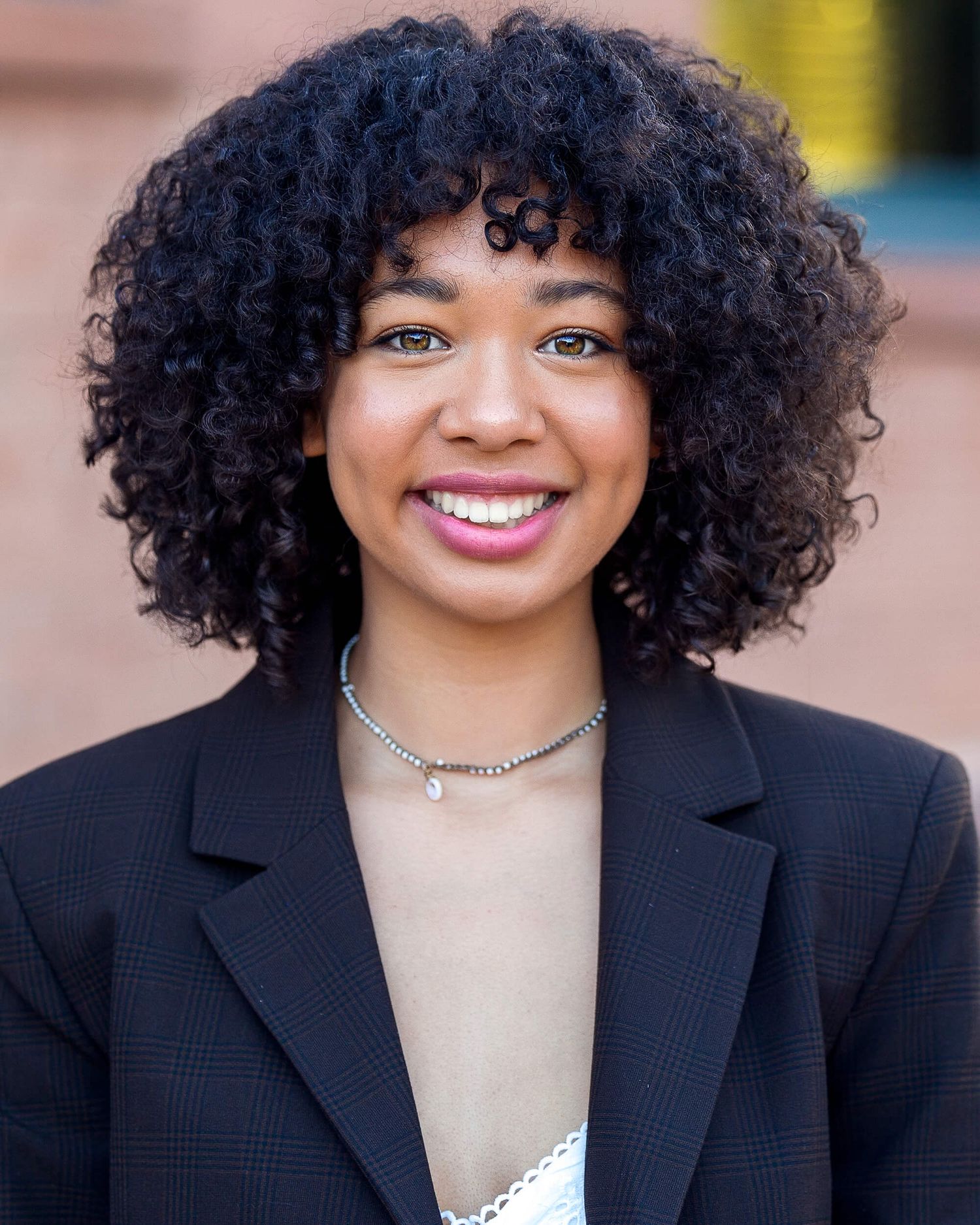 Young woman with curly black hair smiling in a black blazer with a silver necklace outdoors.