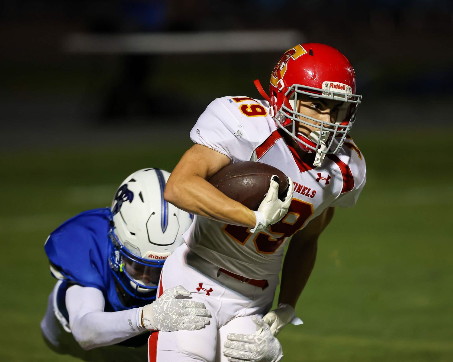 High school football player in red uniform carries the ball while being tackled at night.