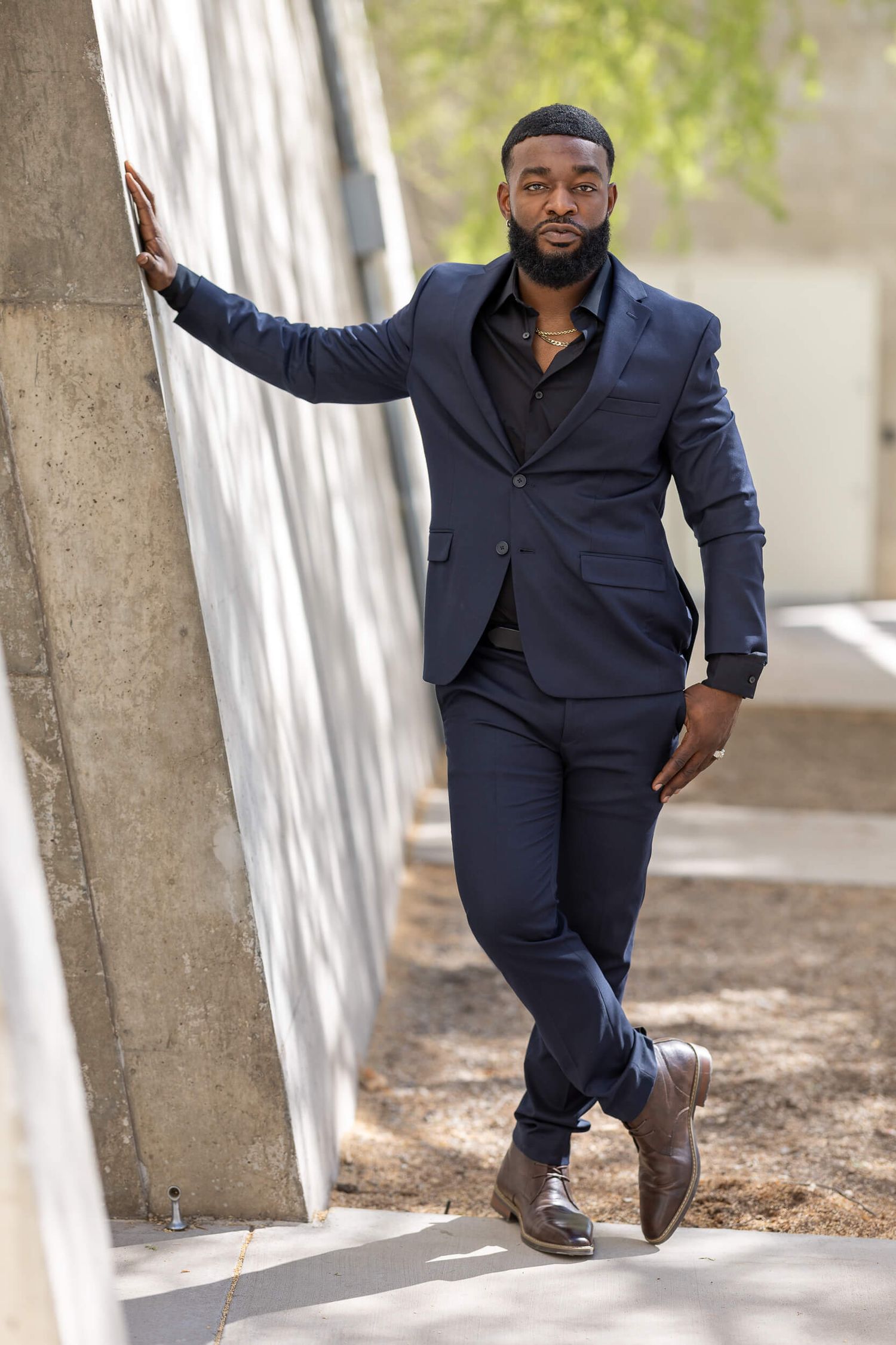 A stylish Black man in a navy suit leans against a concrete wall outdoors wearing brown dress shoes.
