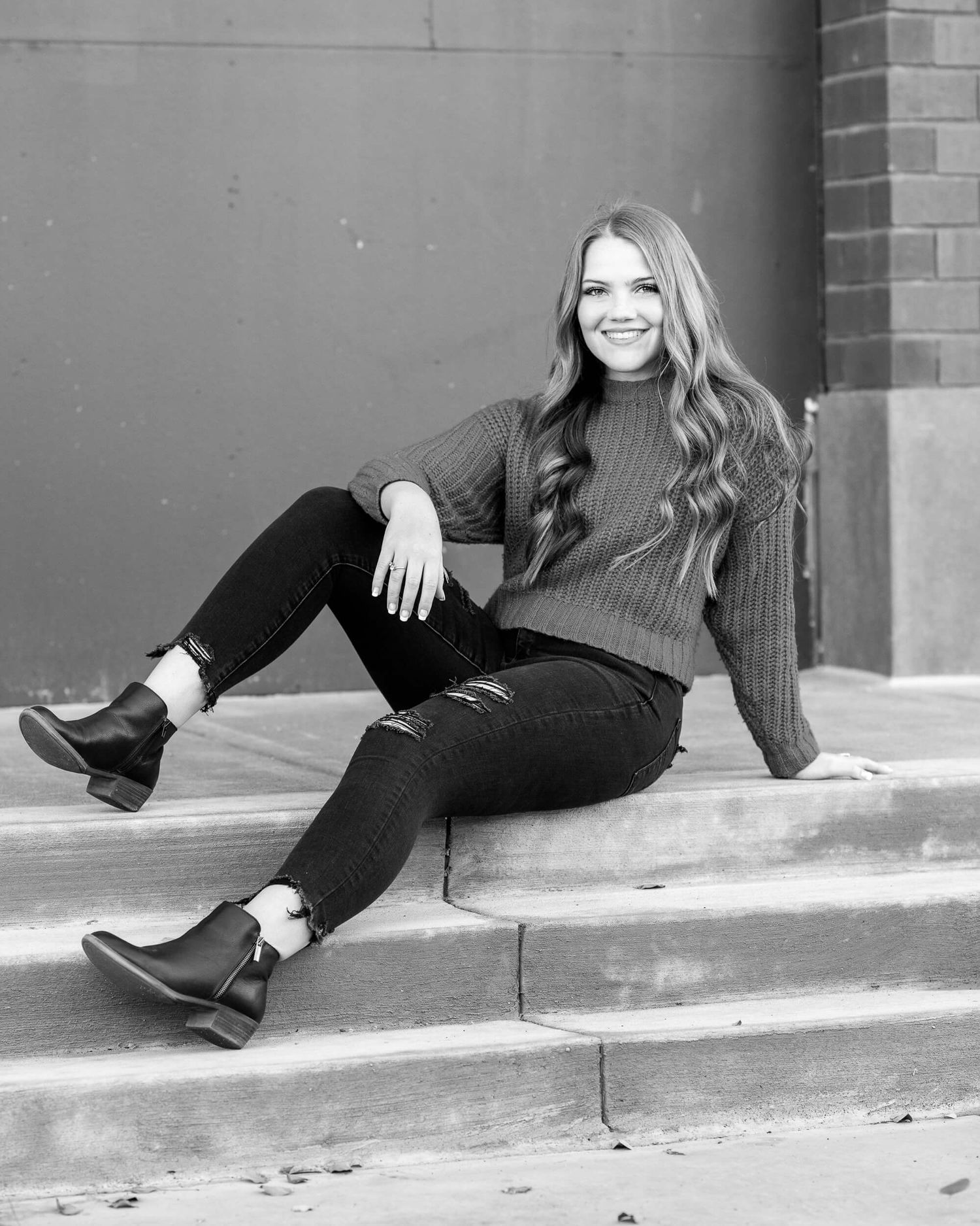 Smiling young woman in black jeans and sweater sits on outdoor steps in a black and white photo.