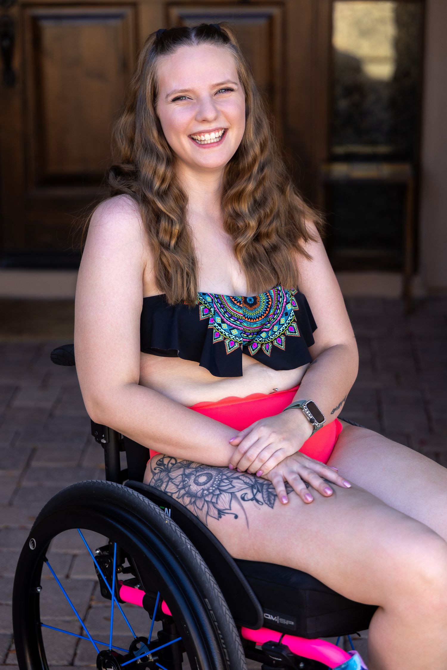 Smiling young woman with tattoos sitting in a pink and black wheelchair, wearing a black bikini top outdoors.