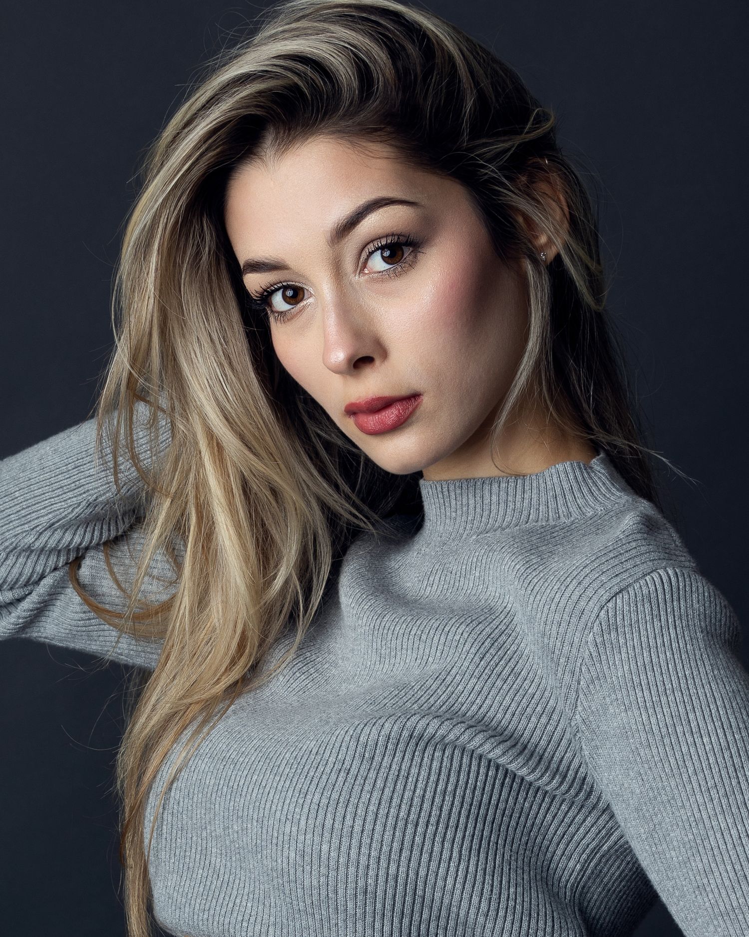 Young woman with blonde highlights in a gray ribbed turtleneck sweater poses against a dark background.