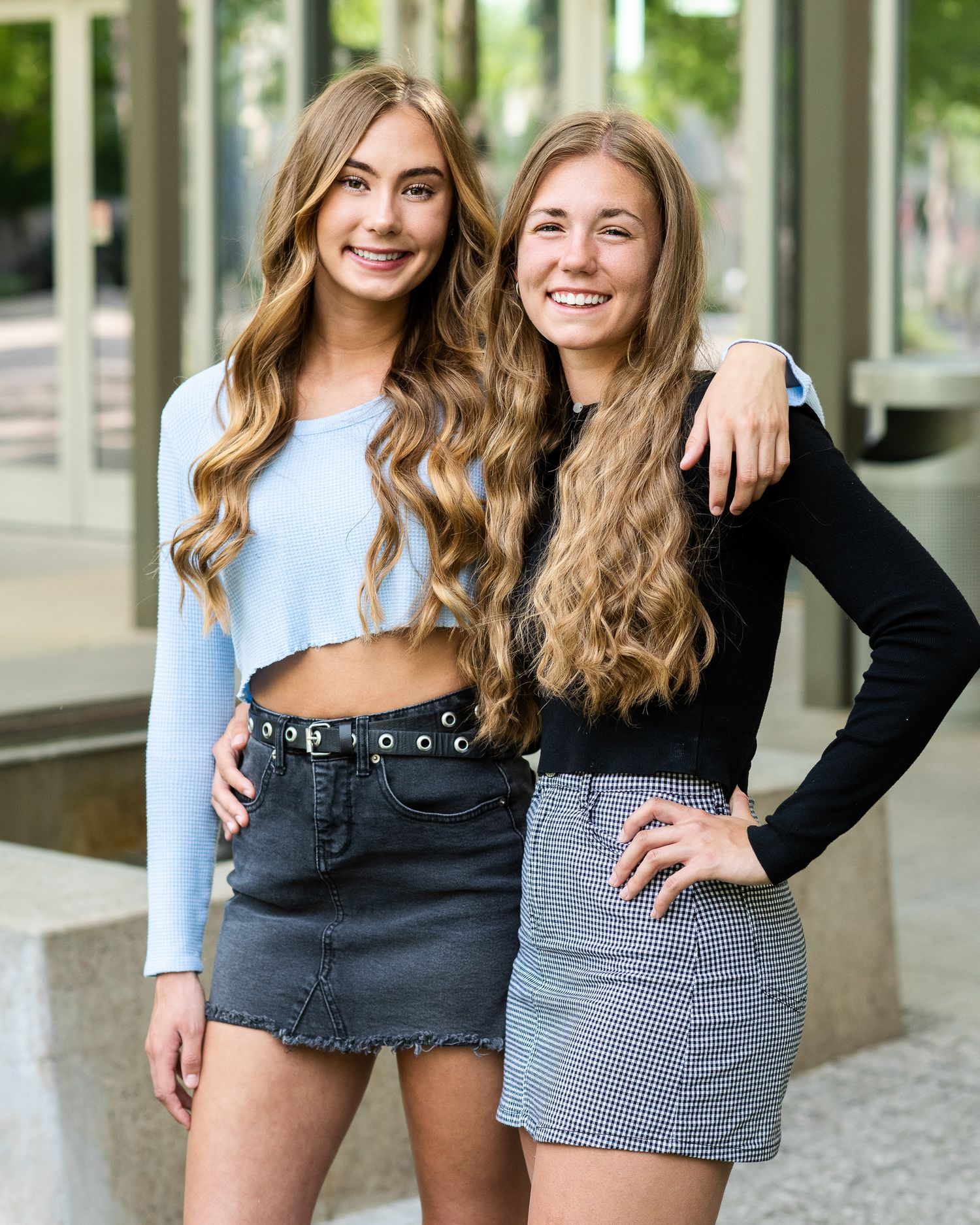 Two smiling teenage girls with long wavy hair wearing mini skirts pose together outdoors.