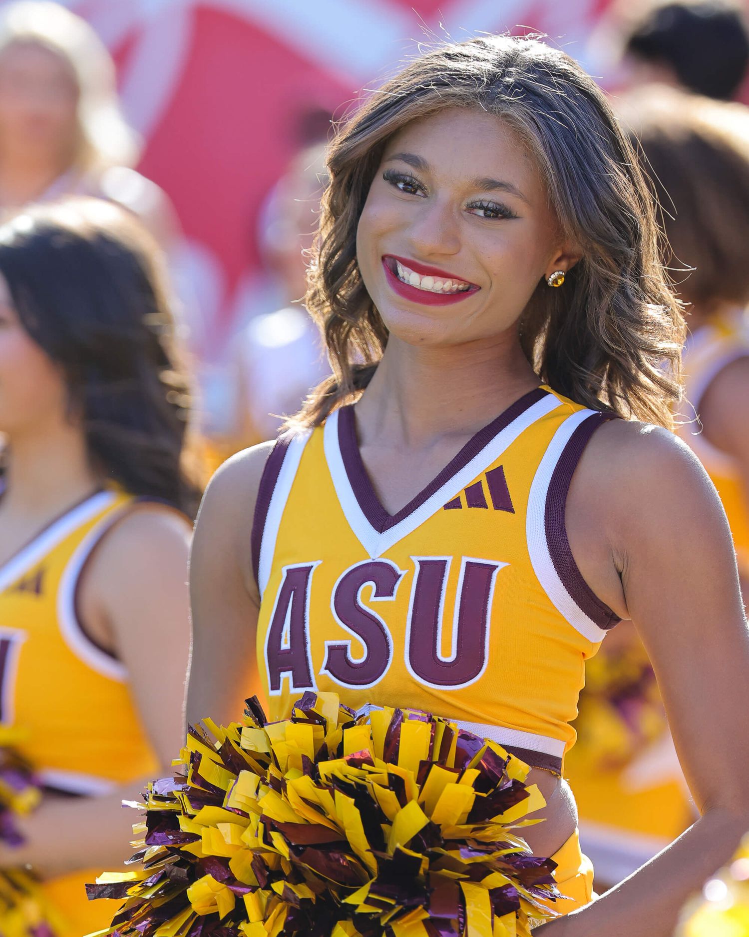 ASU cheerleader smiles in gold and maroon Adidas uniform holding pom-poms at a college football game.