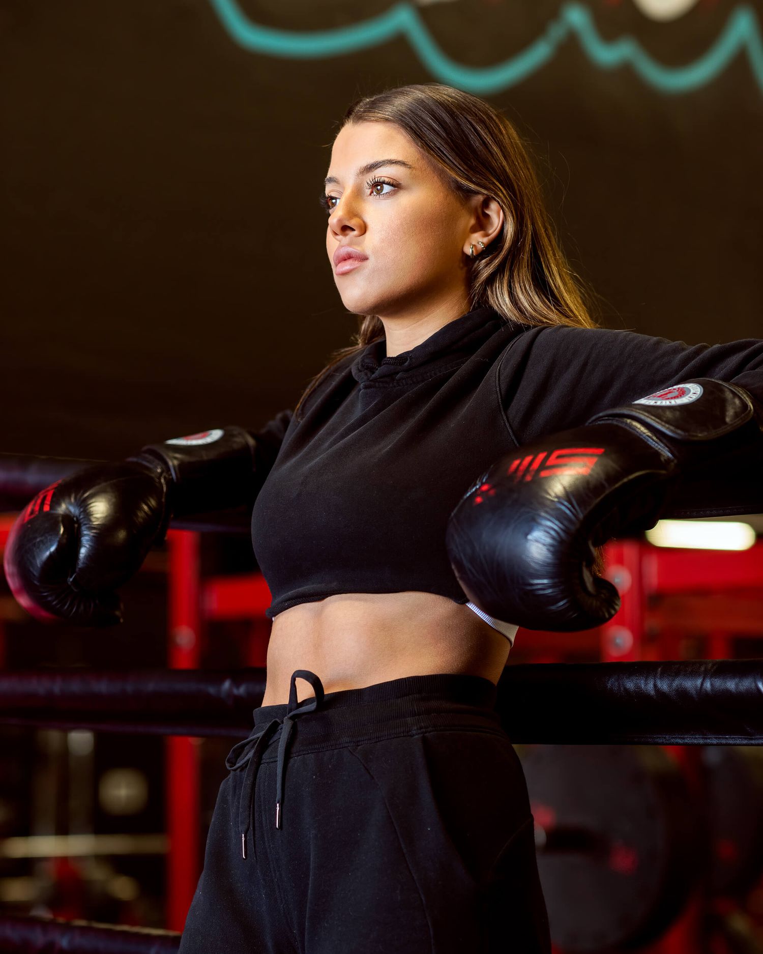 Athletic woman in black crop top and boxing gloves leaning on ropes in a boxing ring.