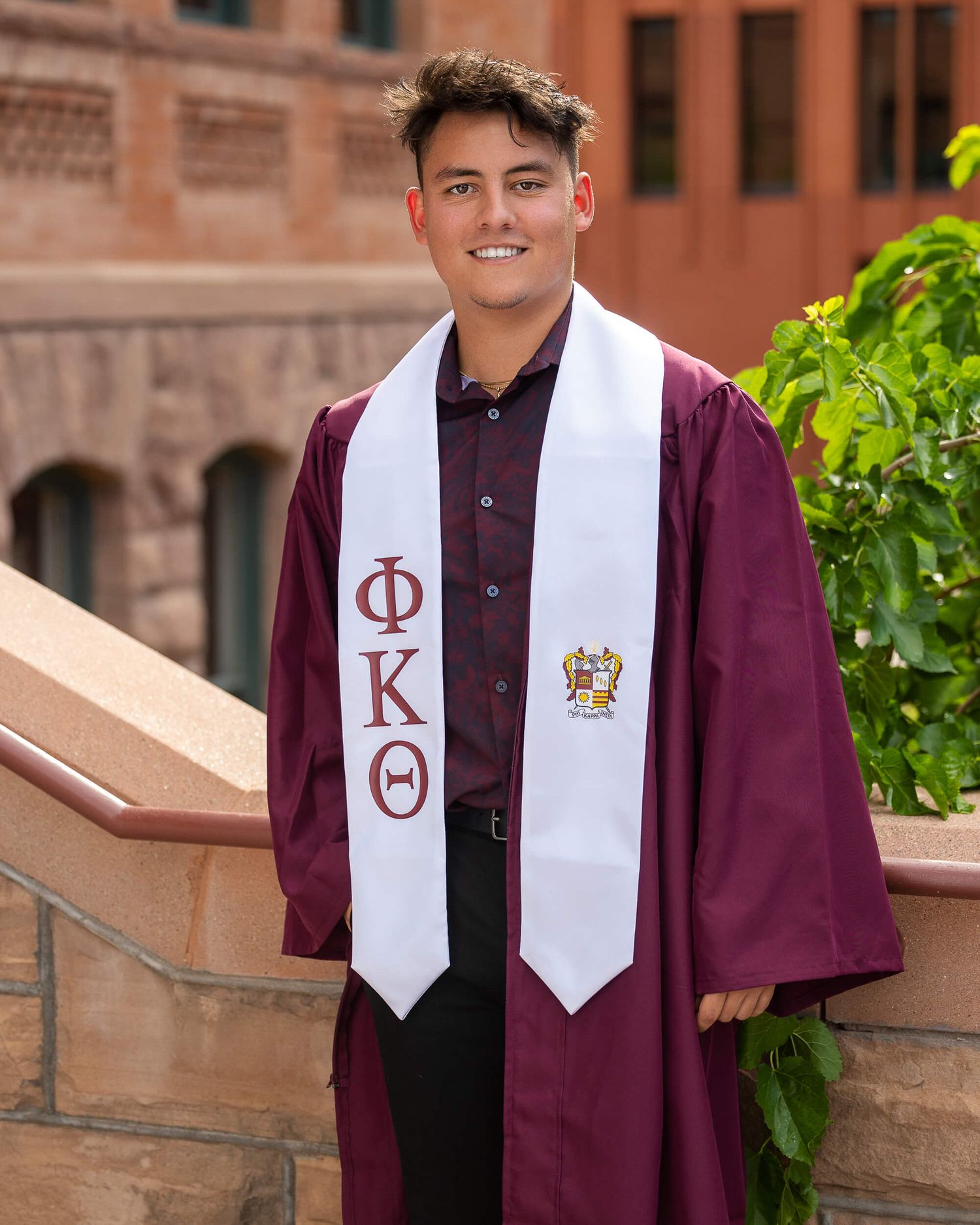 Male graduate smiles in maroon gown with Phi Kappa Theta stole at a brick campus building.