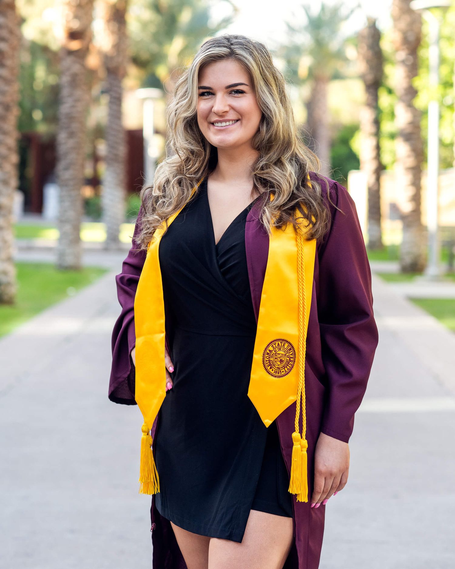 Female graduate in maroon ASU gown and gold stole walking along palm tree-lined path.