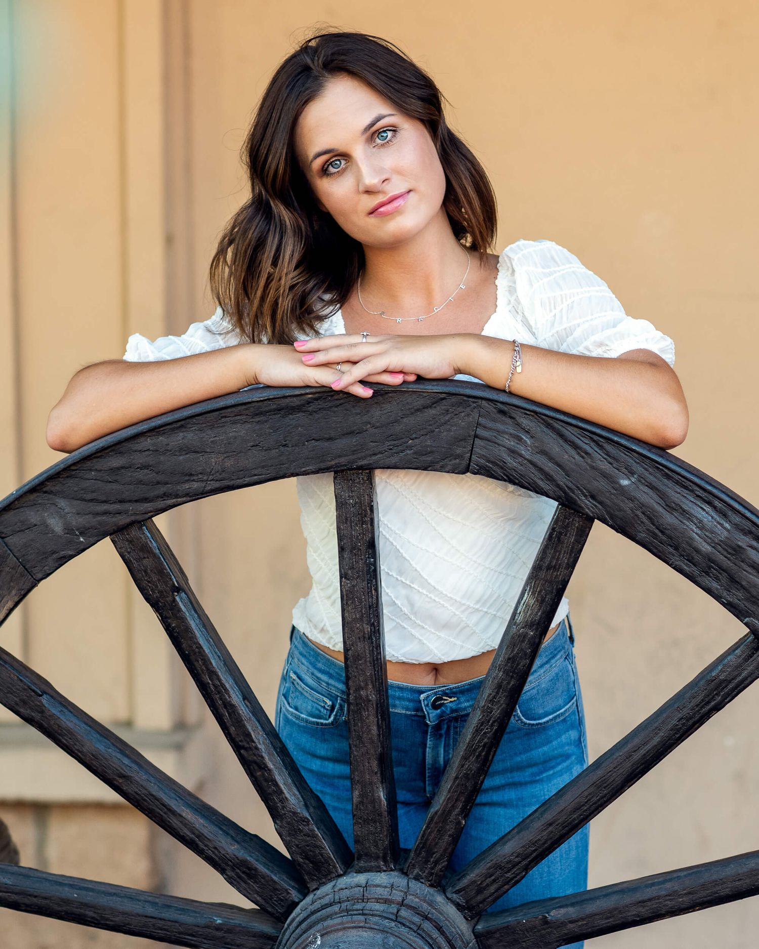 Brunette woman in white top and jeans leans on rustic wooden wagon wheel outdoors.