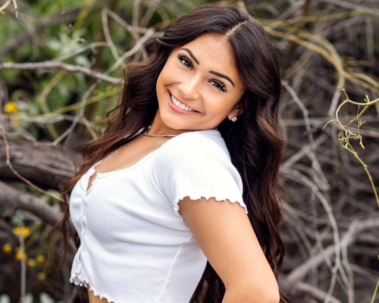 Young woman with long dark wavy hair smiles over her shoulder wearing a white crop top in an outdoor nature setting.