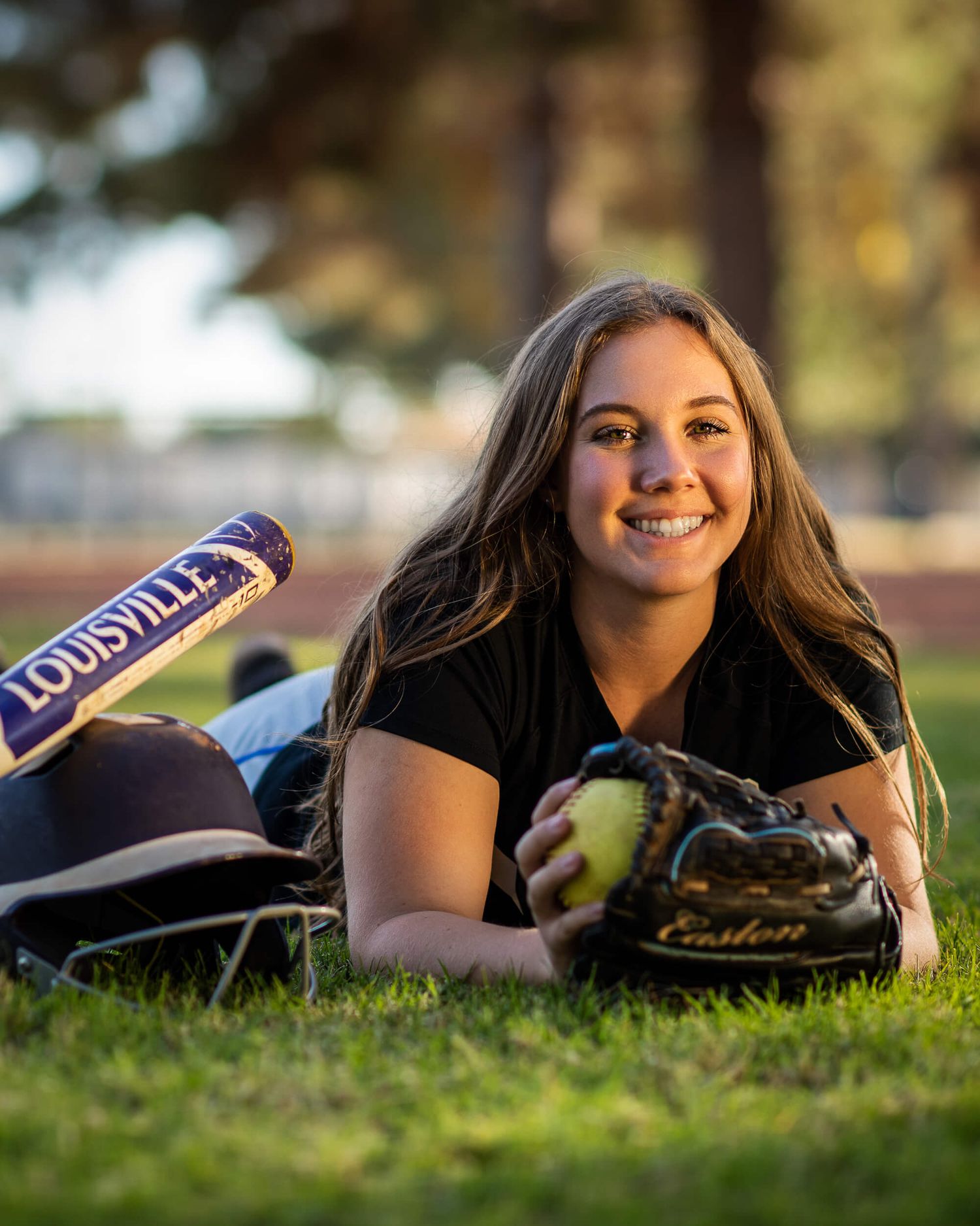 Smiling teen softball player posing with Louisville bat and Easton glove on green grass field.