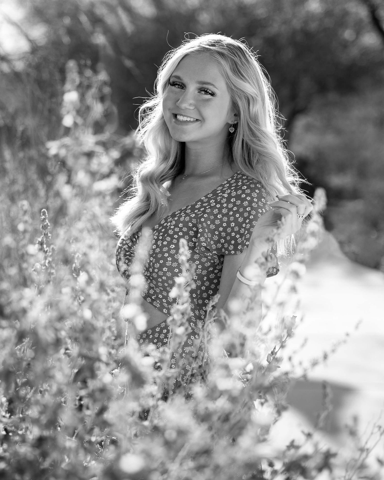 Smiling blonde woman in floral dress surrounded by wildflowers in a black and white outdoor portrait photo.