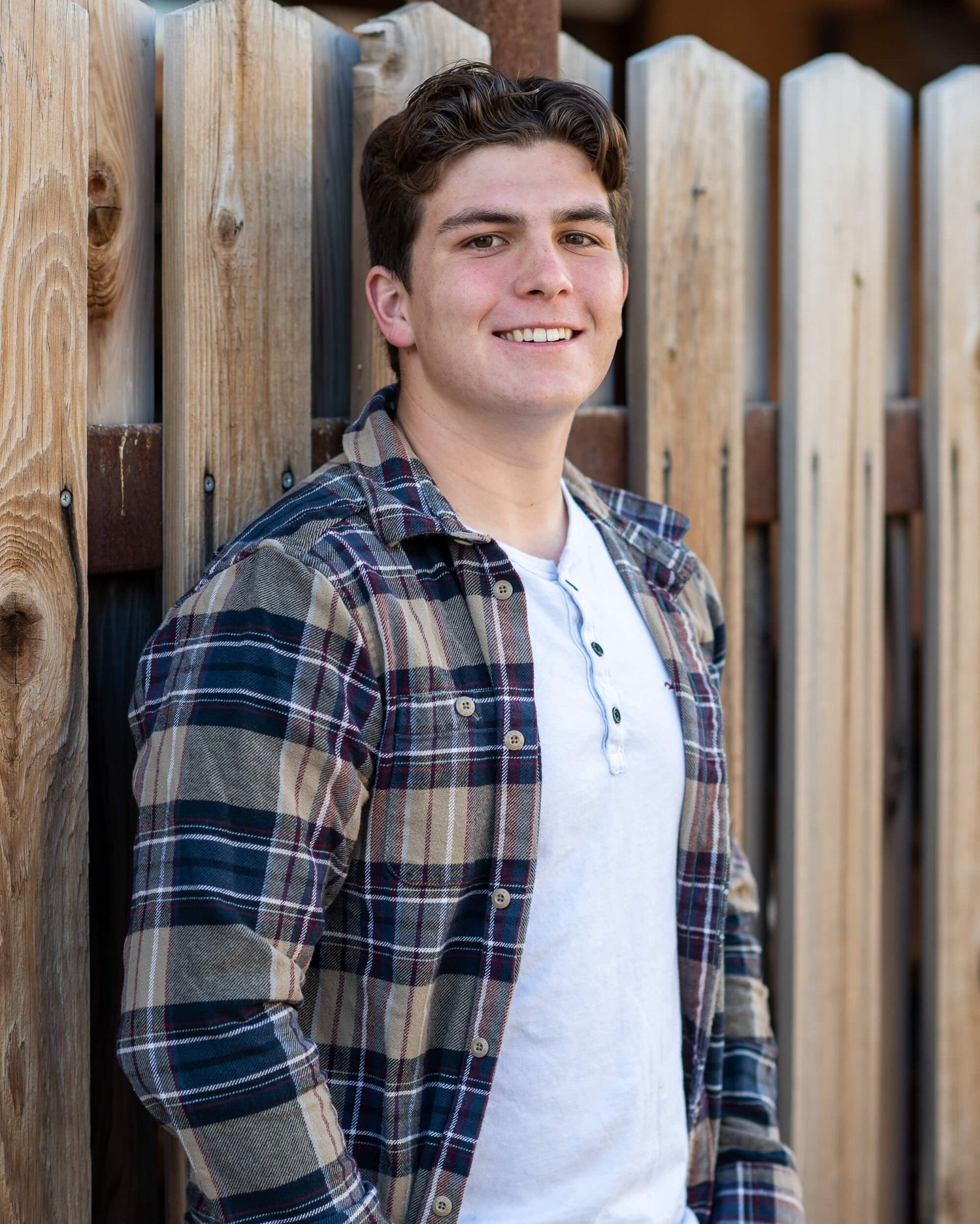 Smiling young man in plaid flannel shirt leaning against wooden fence outdoors.