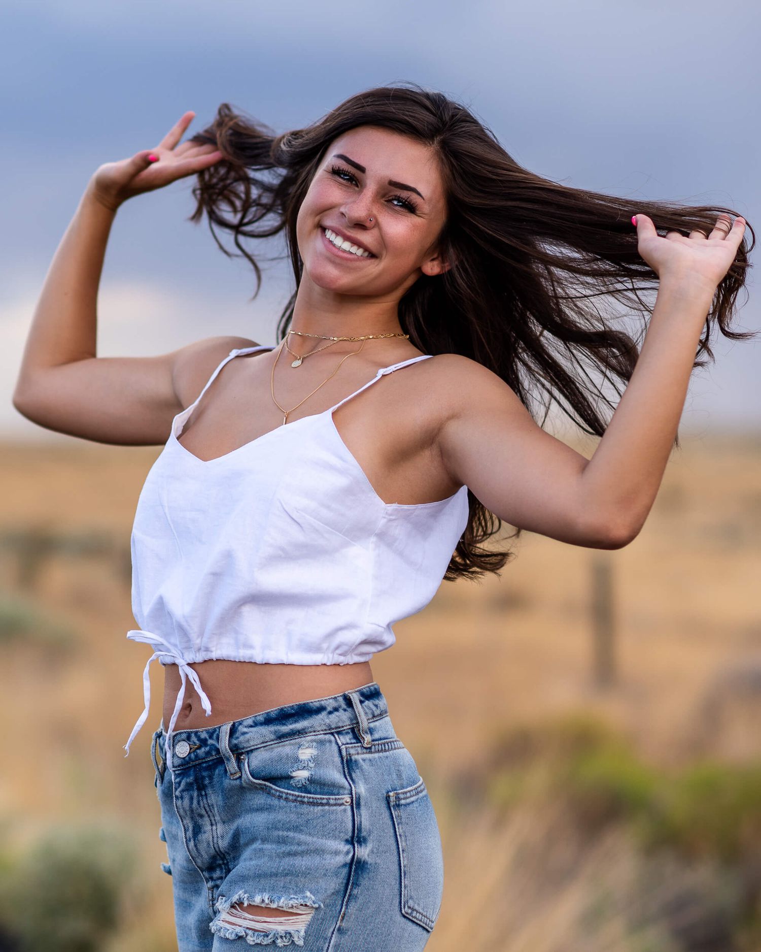 Smiling brunette teen girl senior photo in white crop top and jeans in open field.