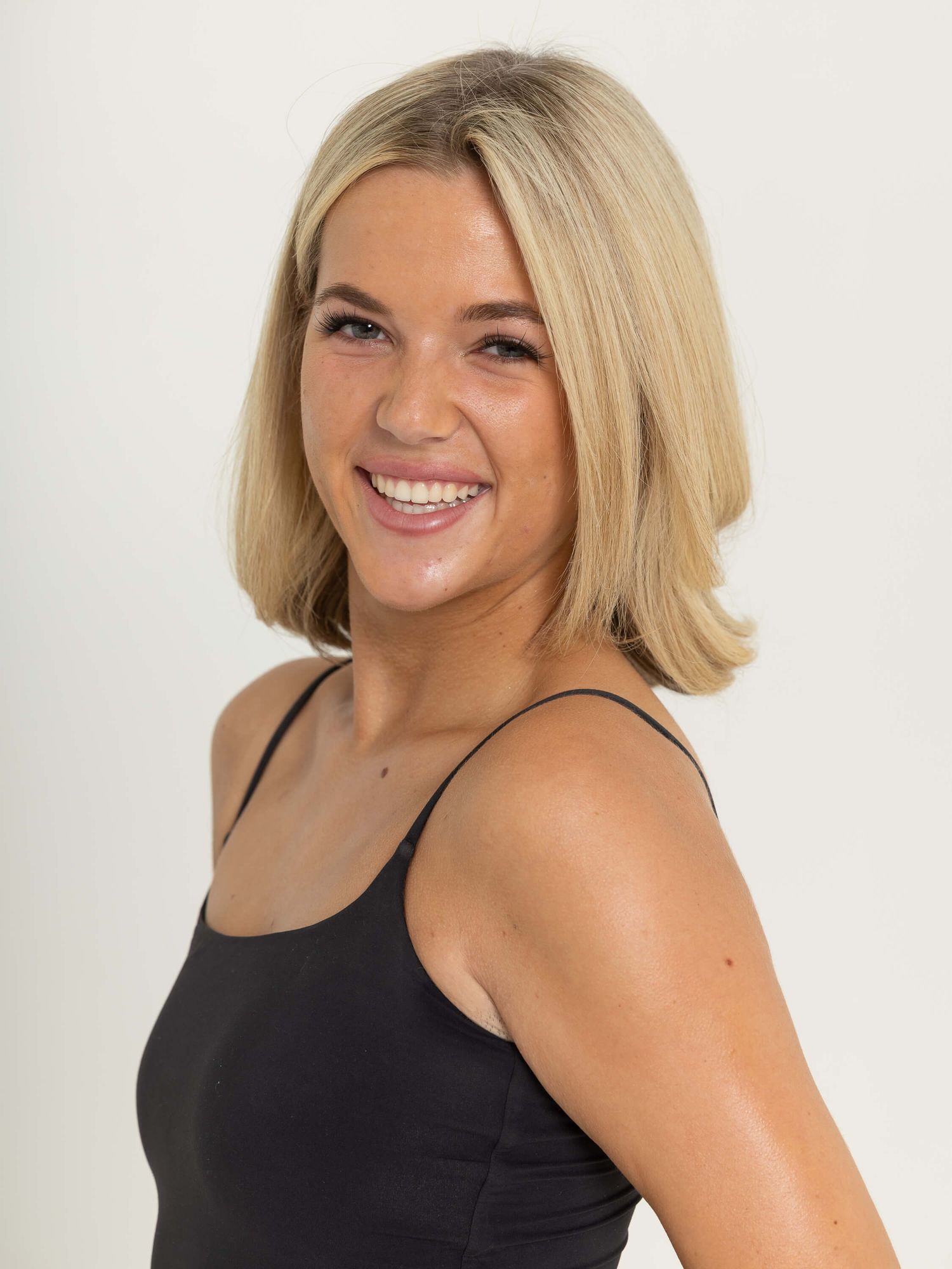 Smiling young woman with blonde bob haircut wearing a black tank top against a white background.