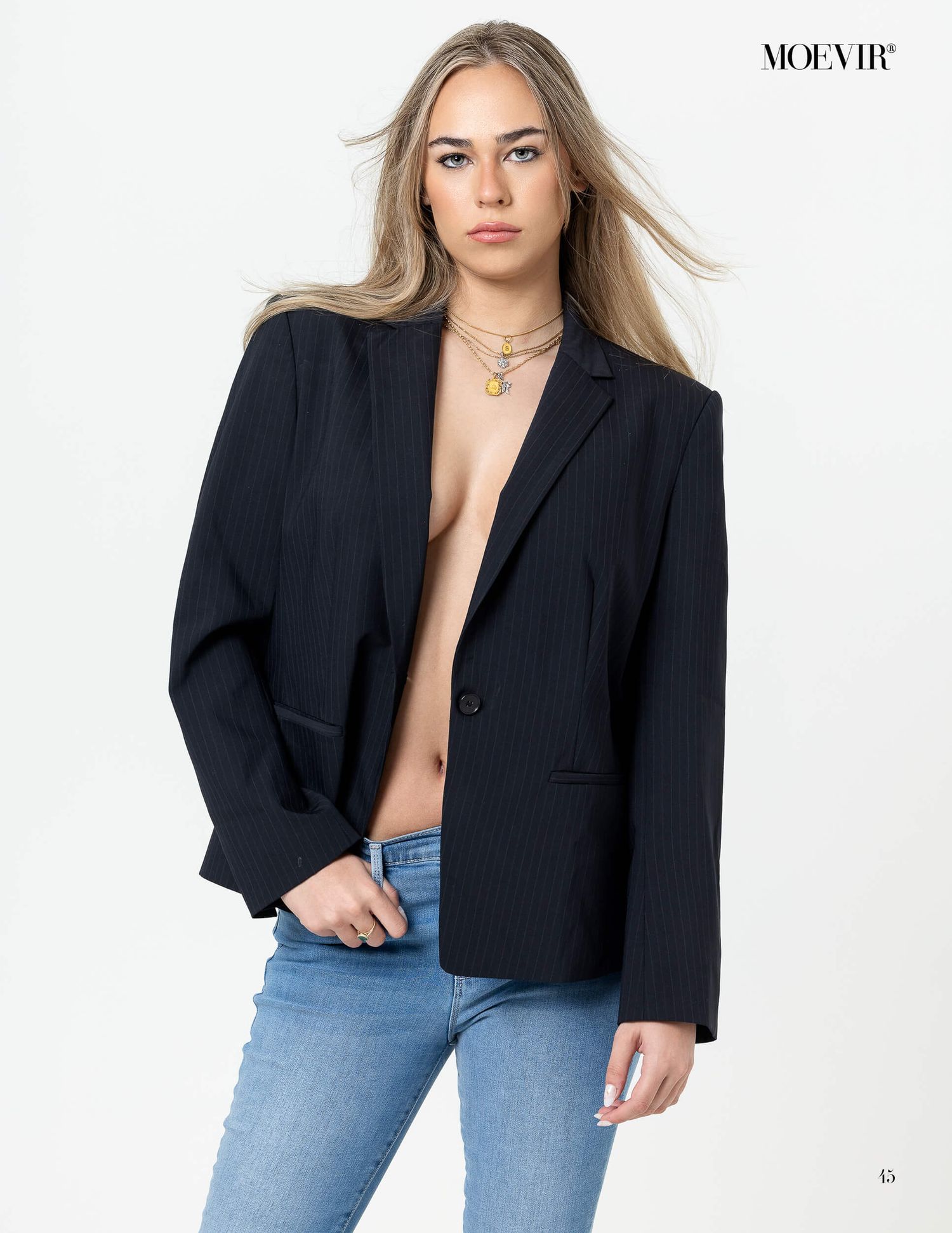 Blonde model poses in open black blazer with layered gold necklaces and blue jeans for Moevir Magazine.