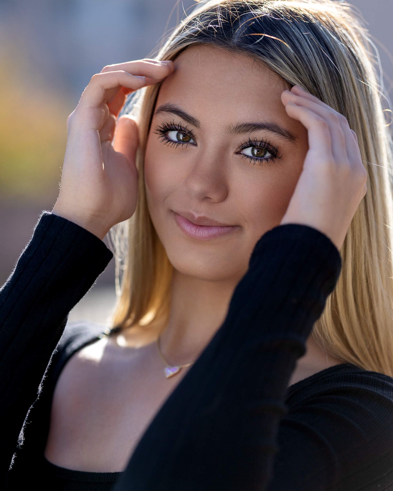 Young blonde woman with hazel eyes holding hands to head smiling outdoors in natural light.