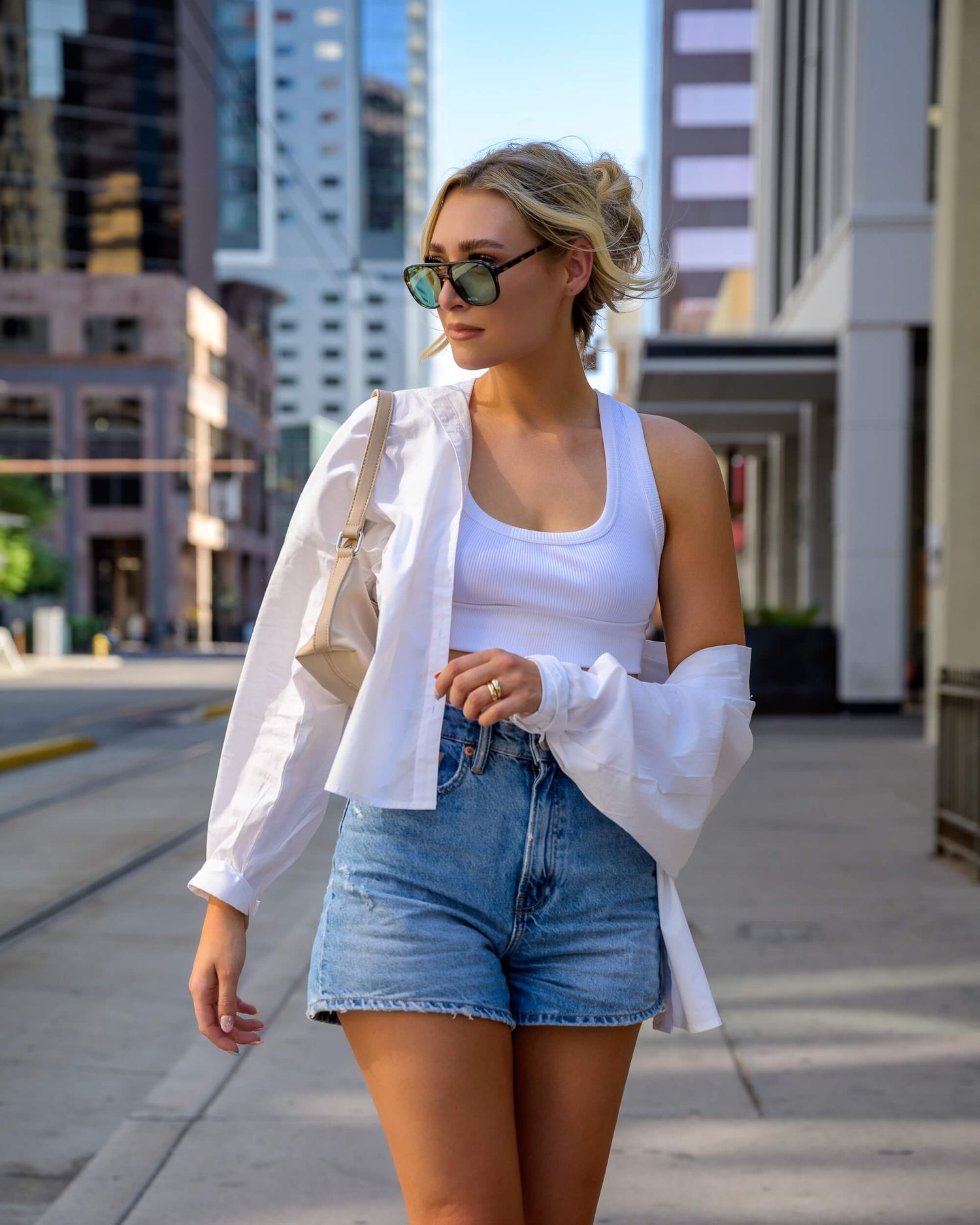 Blonde woman in white tank, denim shorts, and open white shirt walking city street.