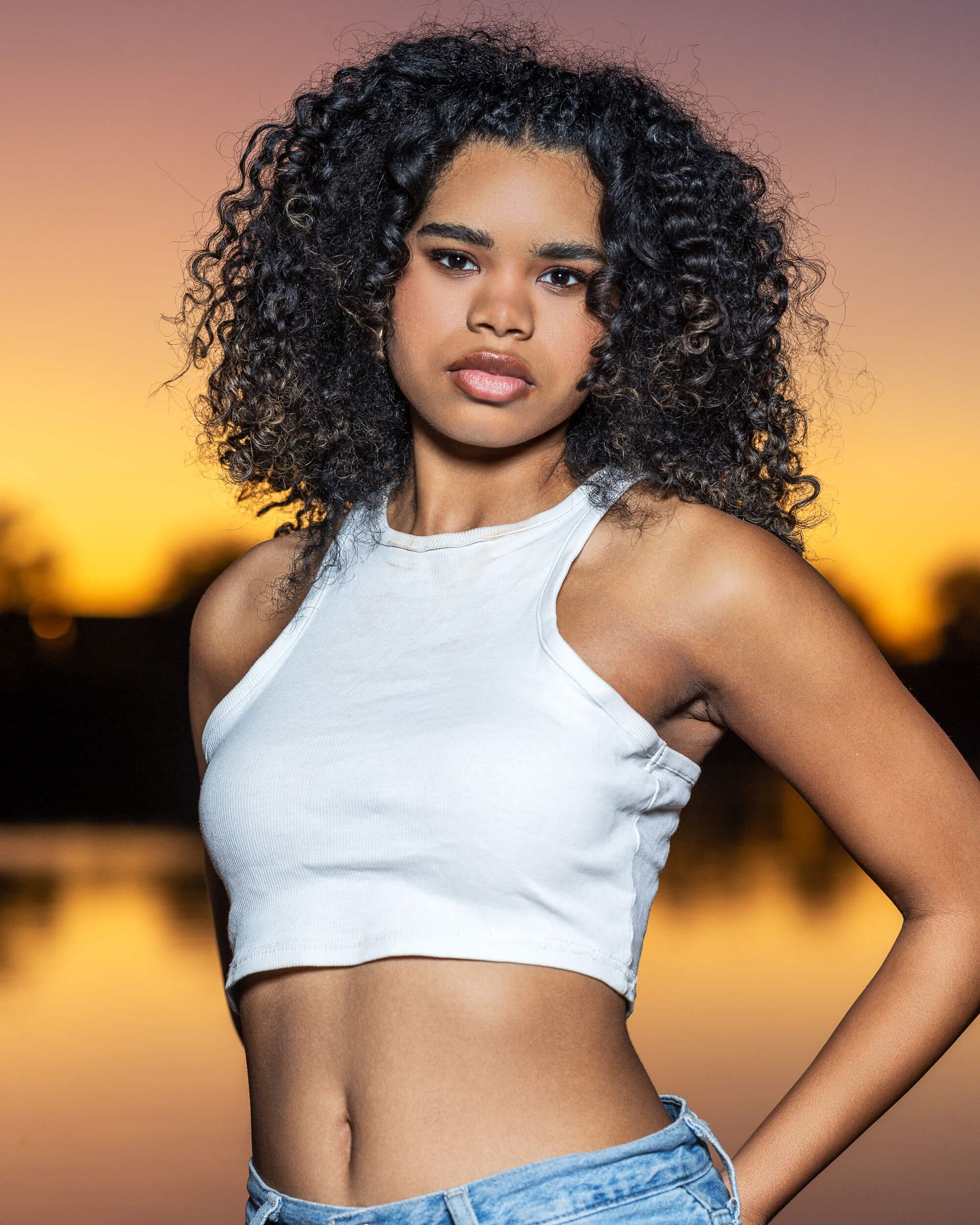 Young woman with curly hair poses confidently in white crop top and jeans against a golden sunset backdrop.