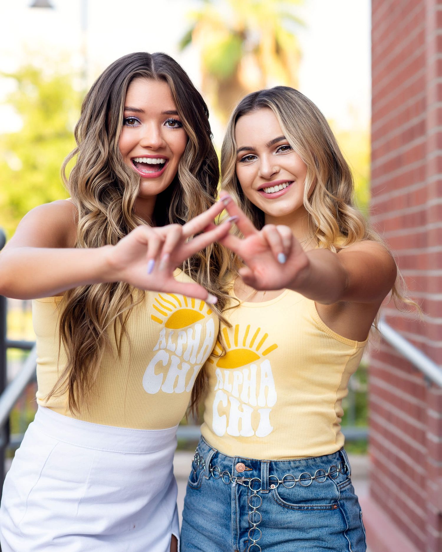 Two smiling women in matching yellow Alpha Chi sorority tops making heart shapes with their hands outdoors.