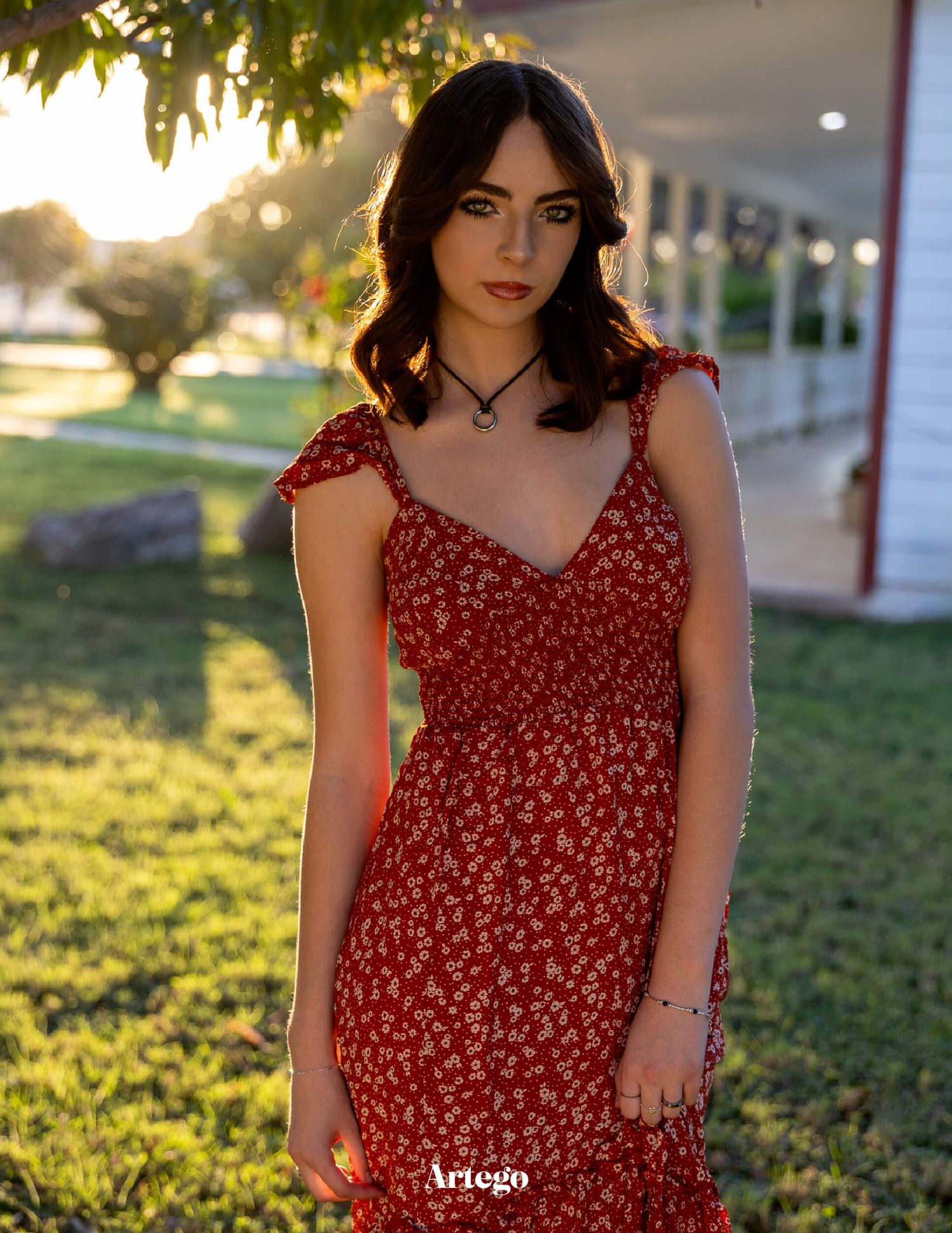 Young woman in red floral sundress poses outdoors in golden hour sunlight near a white building.