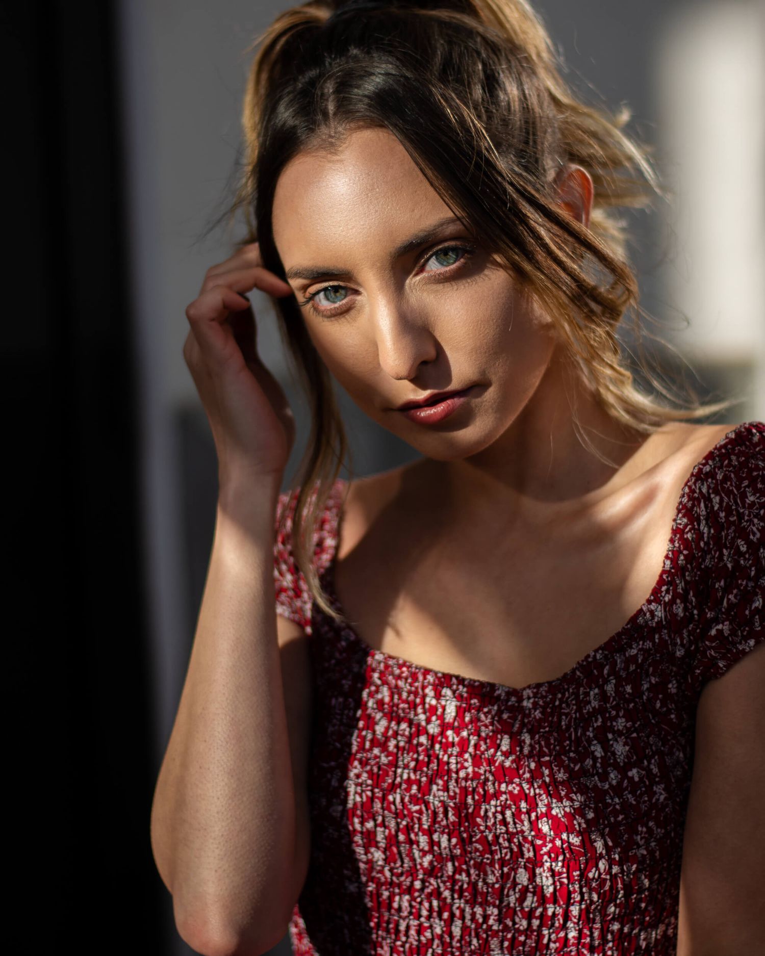 Young woman in red floral dress poses in dramatic sunlight with hand near face.