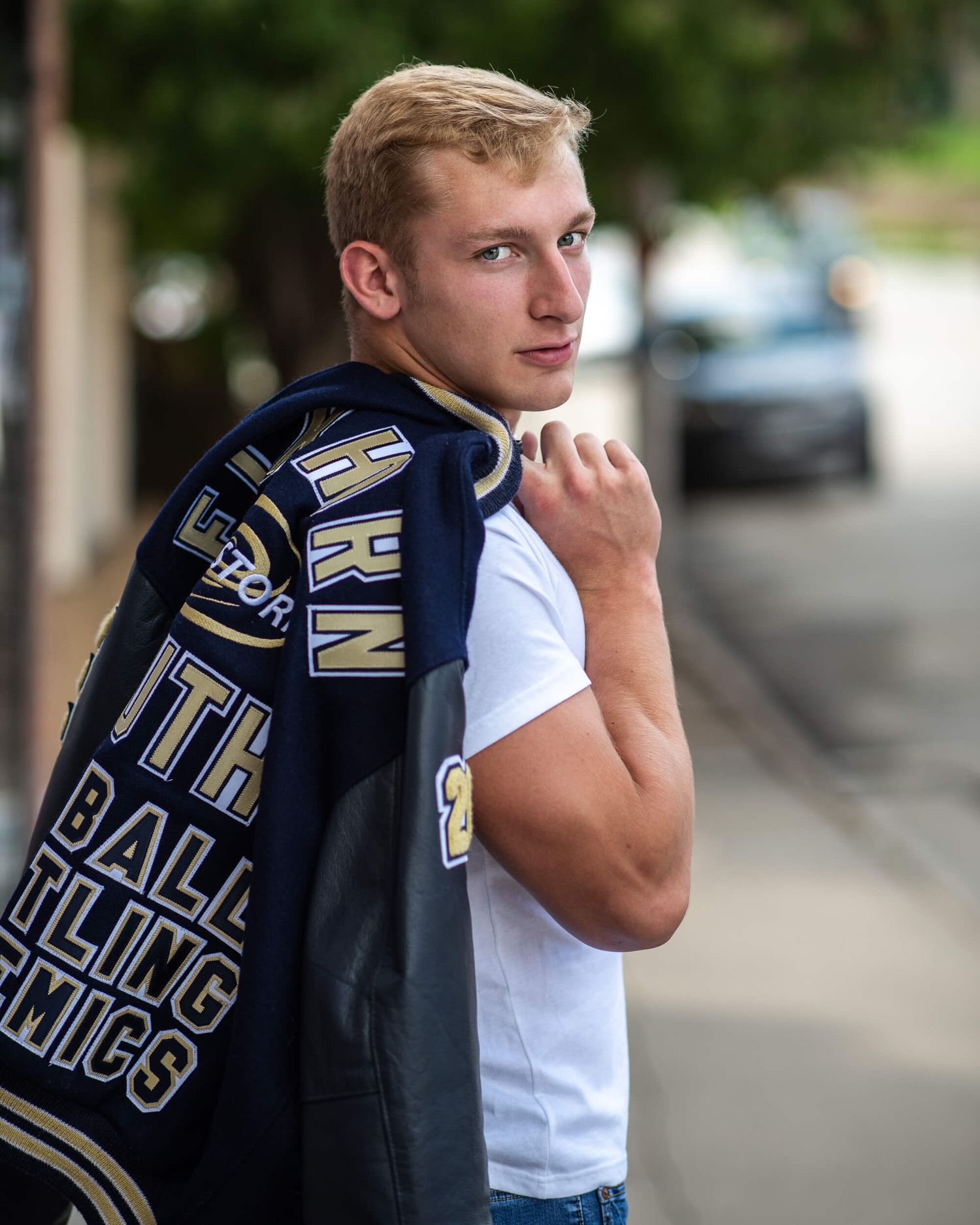 Athletic teenage boy in white tee holding varsity jacket over shoulder outdoors.