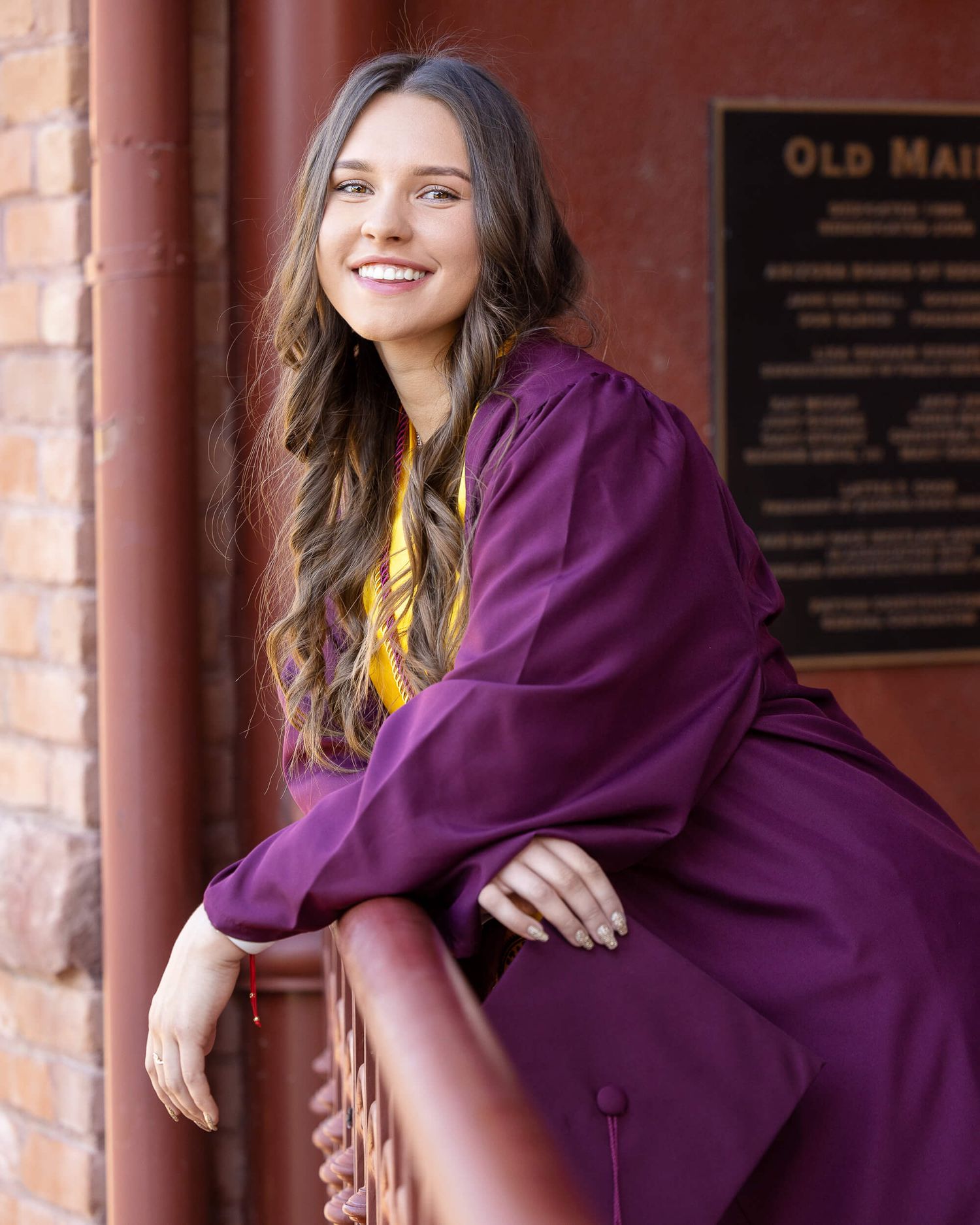 Young woman smiling in maroon graduation gown leaning on railing near Old Main building.