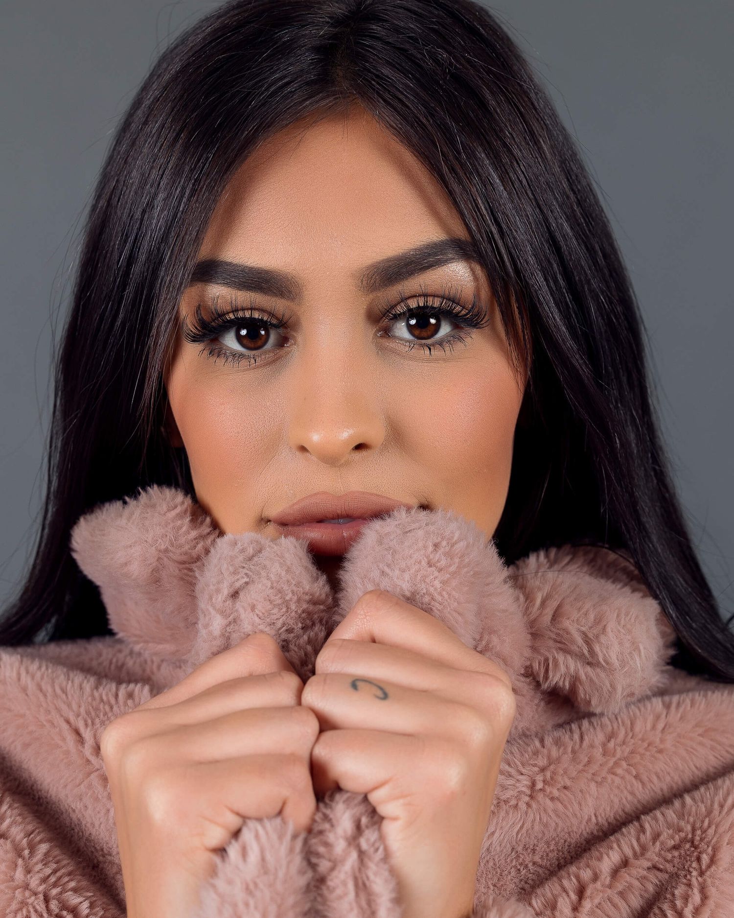 Young woman with dark hair and bold makeup poses in pink fluffy coat.