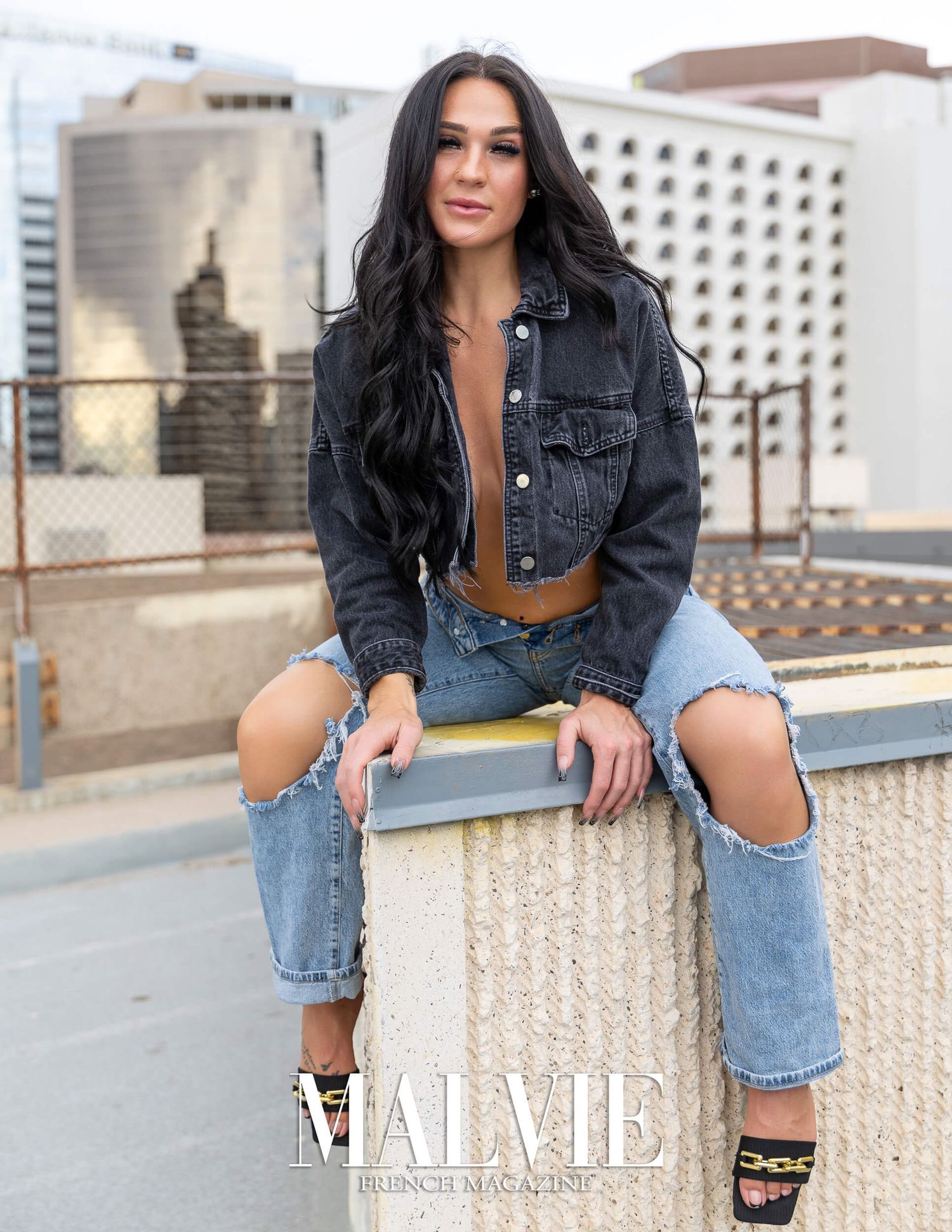 Dark-haired woman in ripped jeans and denim jacket posing outdoors for Malvie Magazine.