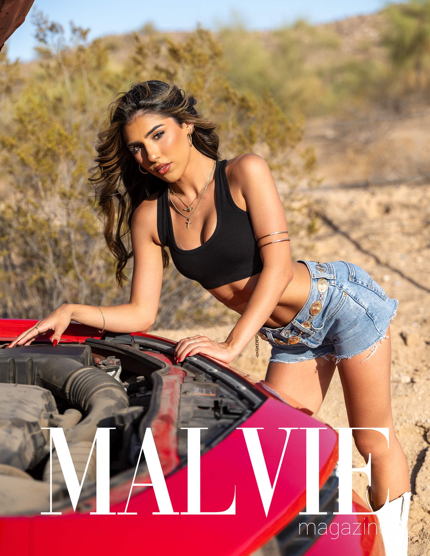 Model in black crop top and denim shorts leans over red convertible for MALVIE Magazine desert shoot.