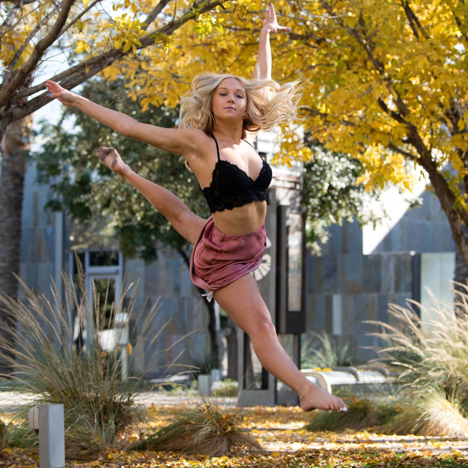 Blonde female dancer leaps gracefully outdoors among autumn trees in a black crop top and pink skirt.