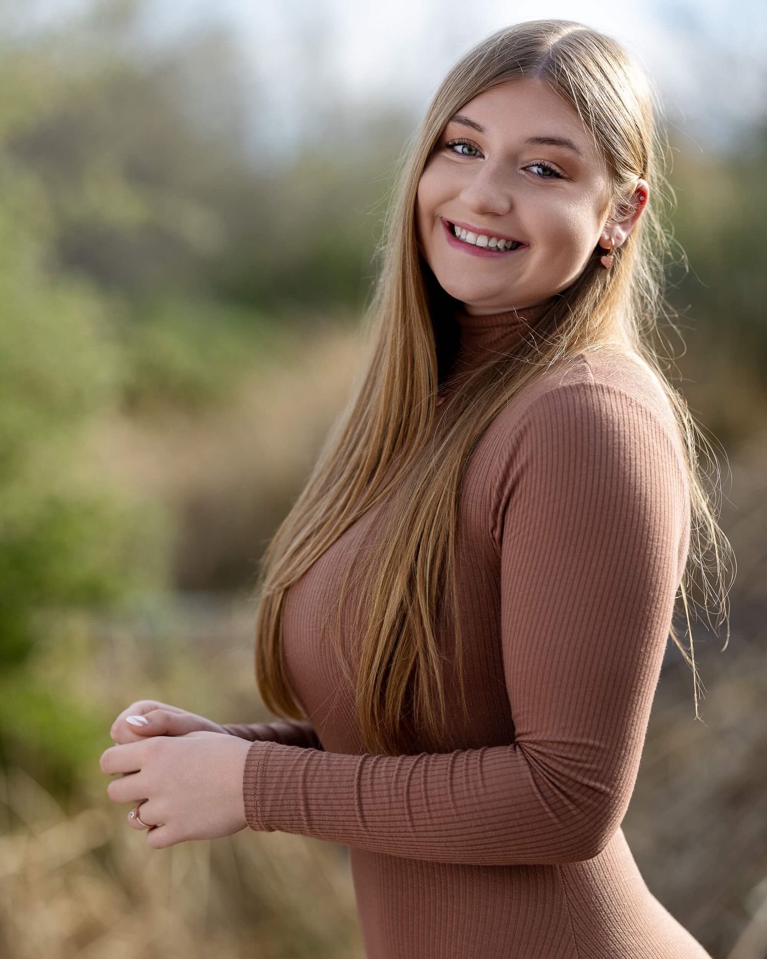 Smiling young woman with long blonde hair in brown turtleneck outdoors.