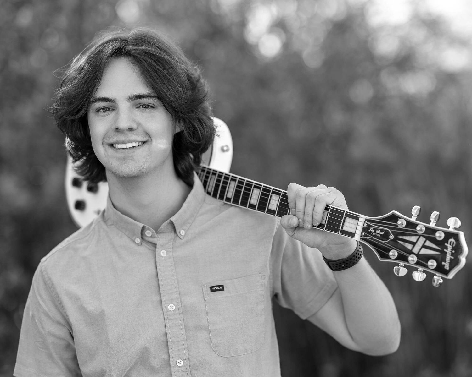 Smiling teenage boy with dark hair holds an electric guitar over his shoulder in a black and white outdoor portrait.
