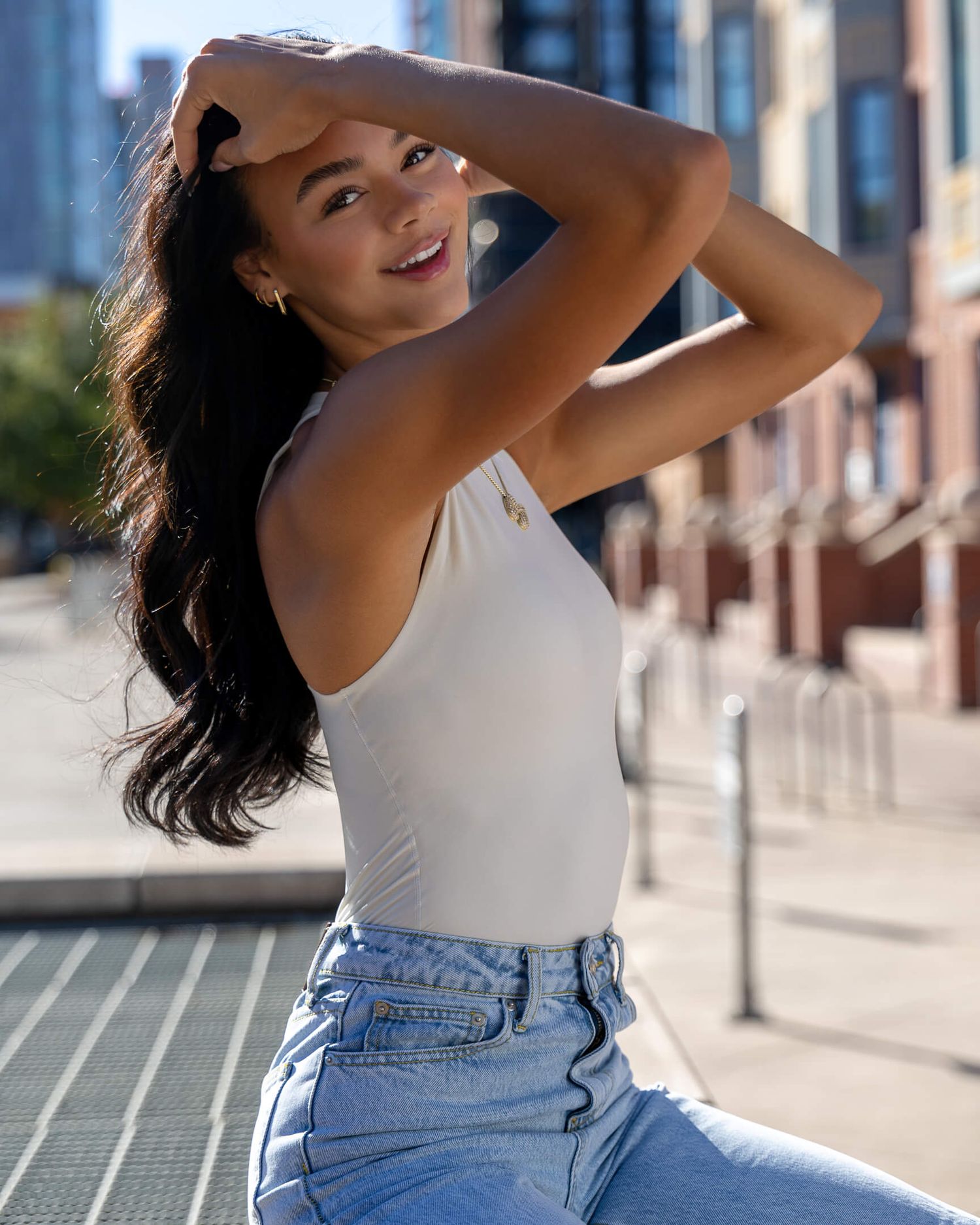 Young woman in white bodysuit and jeans poses outdoors on urban street with buildings in background.