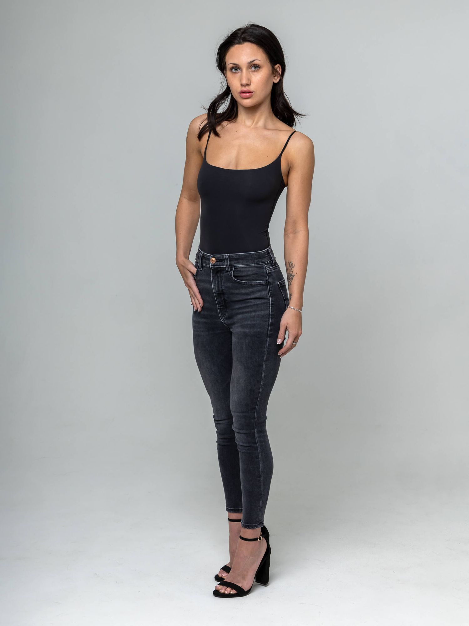 Woman wearing black bodysuit and dark gray skinny jeans with black heeled sandals, posing against gray background.