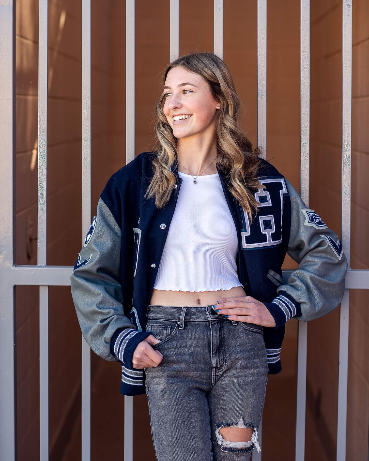 Smiling young woman in navy varsity jacket and ripped jeans posing outdoors against a brown wall.