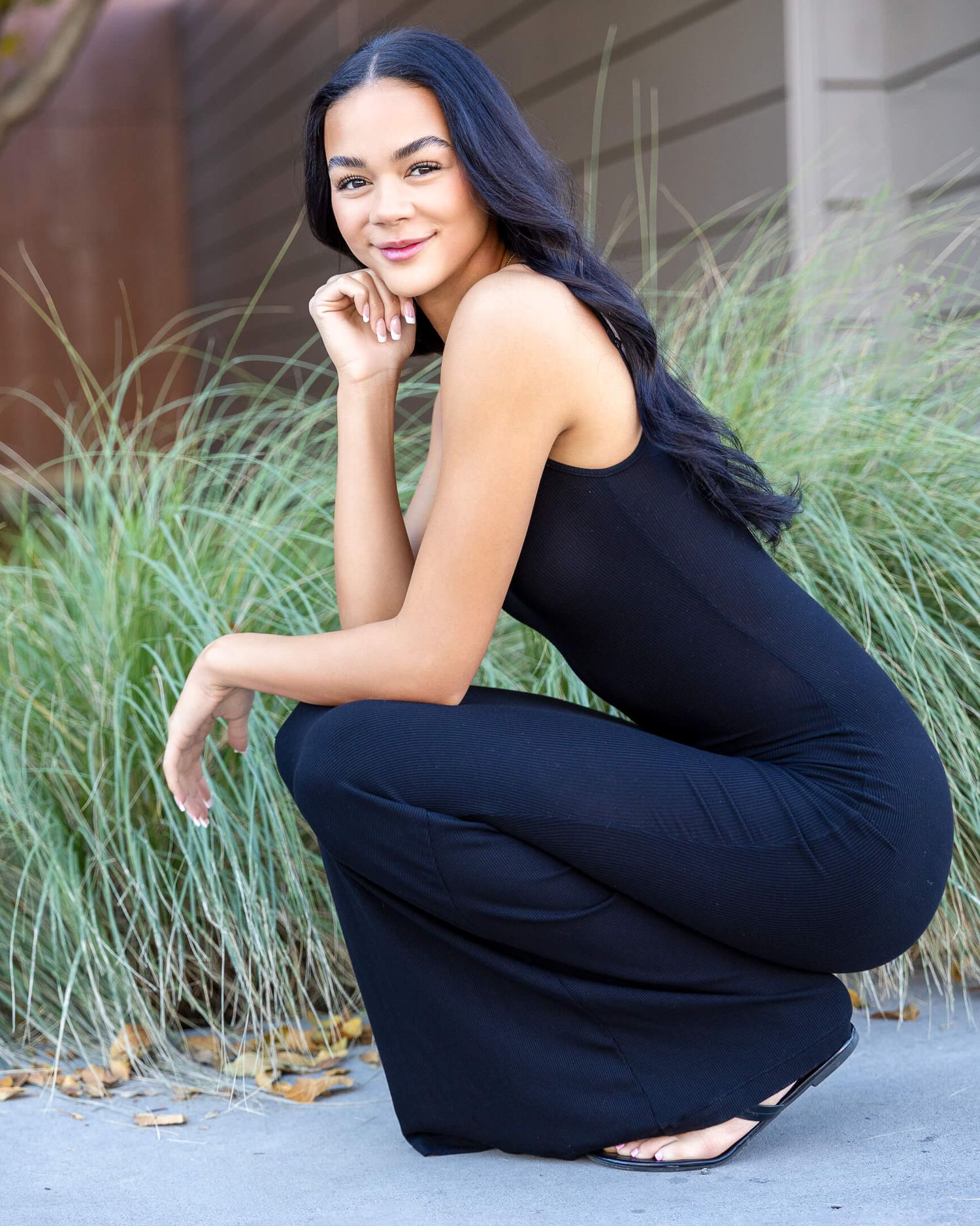 Young woman in black jumpsuit crouching outdoors, smiling, with ornamental grasses in the background.