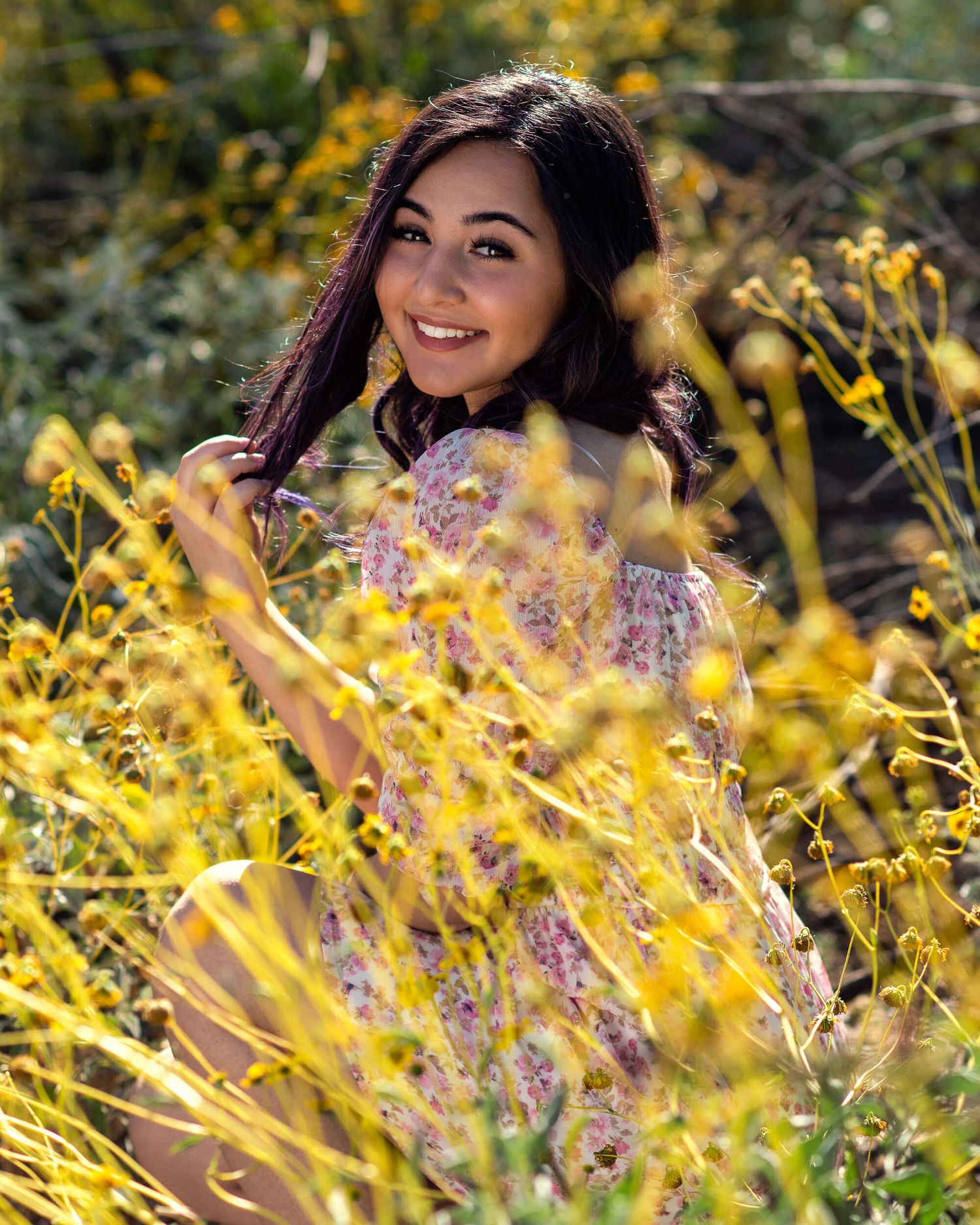 Smiling young woman in floral dress surrounded by vibrant yellow wildflowers in nature.