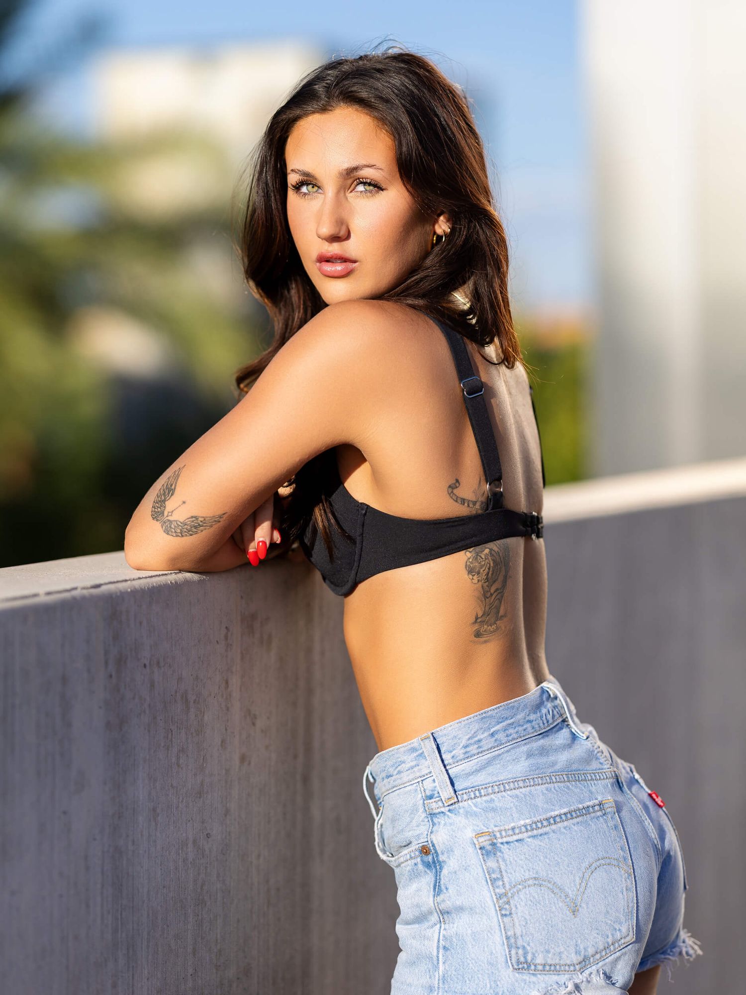 A tattooed woman in a black crop top and Levi's denim shorts poses against a concrete wall outdoors.