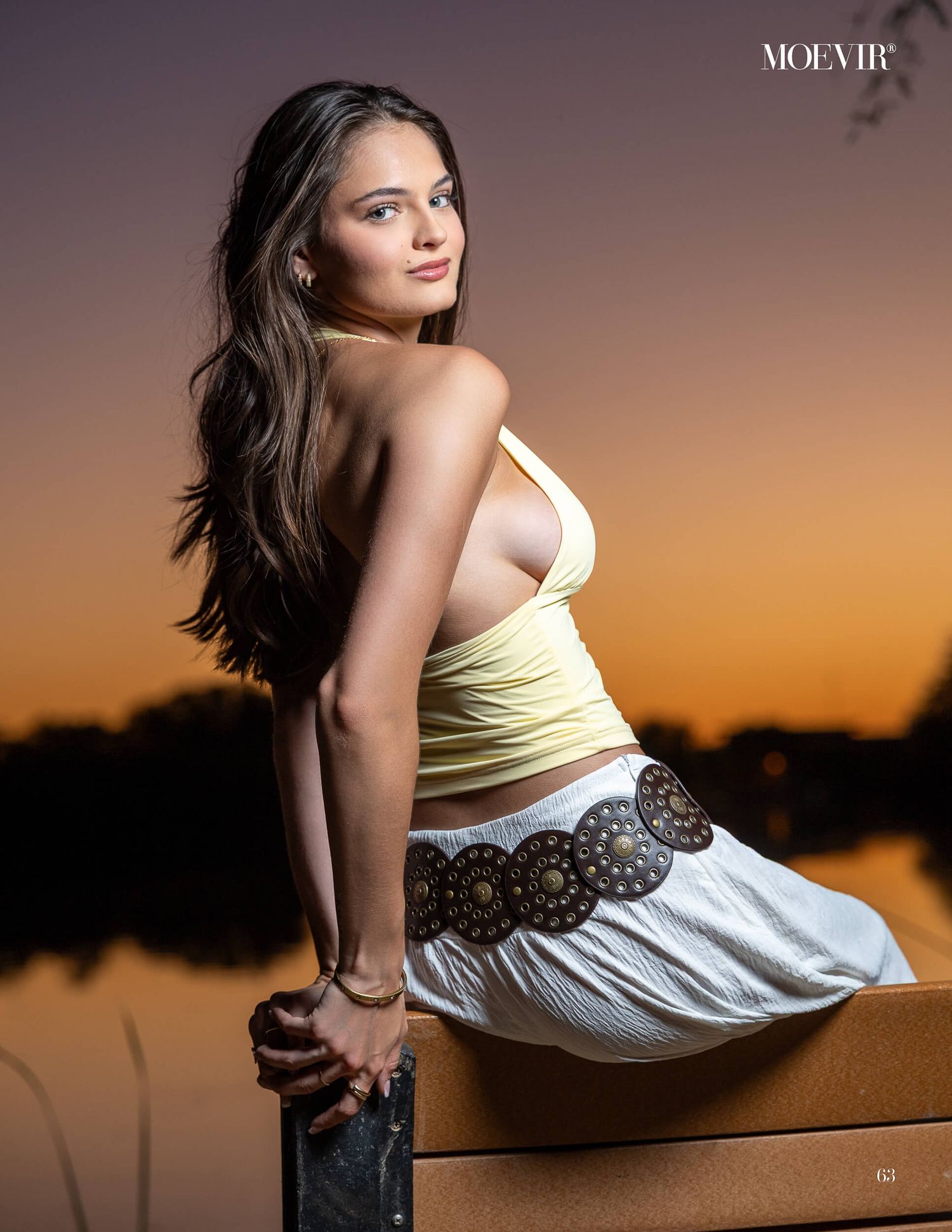 Young woman in yellow crop top and white skirt poses on dock at sunset by a lake.