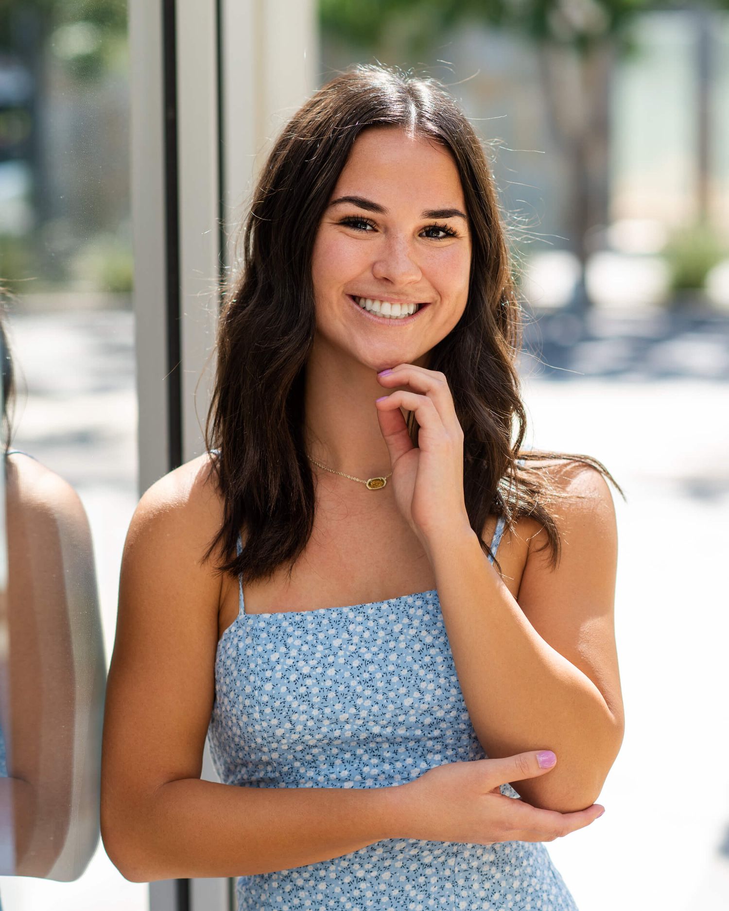 Smiling young woman with dark hair wearing a blue floral sundress, posing outdoors in natural light.