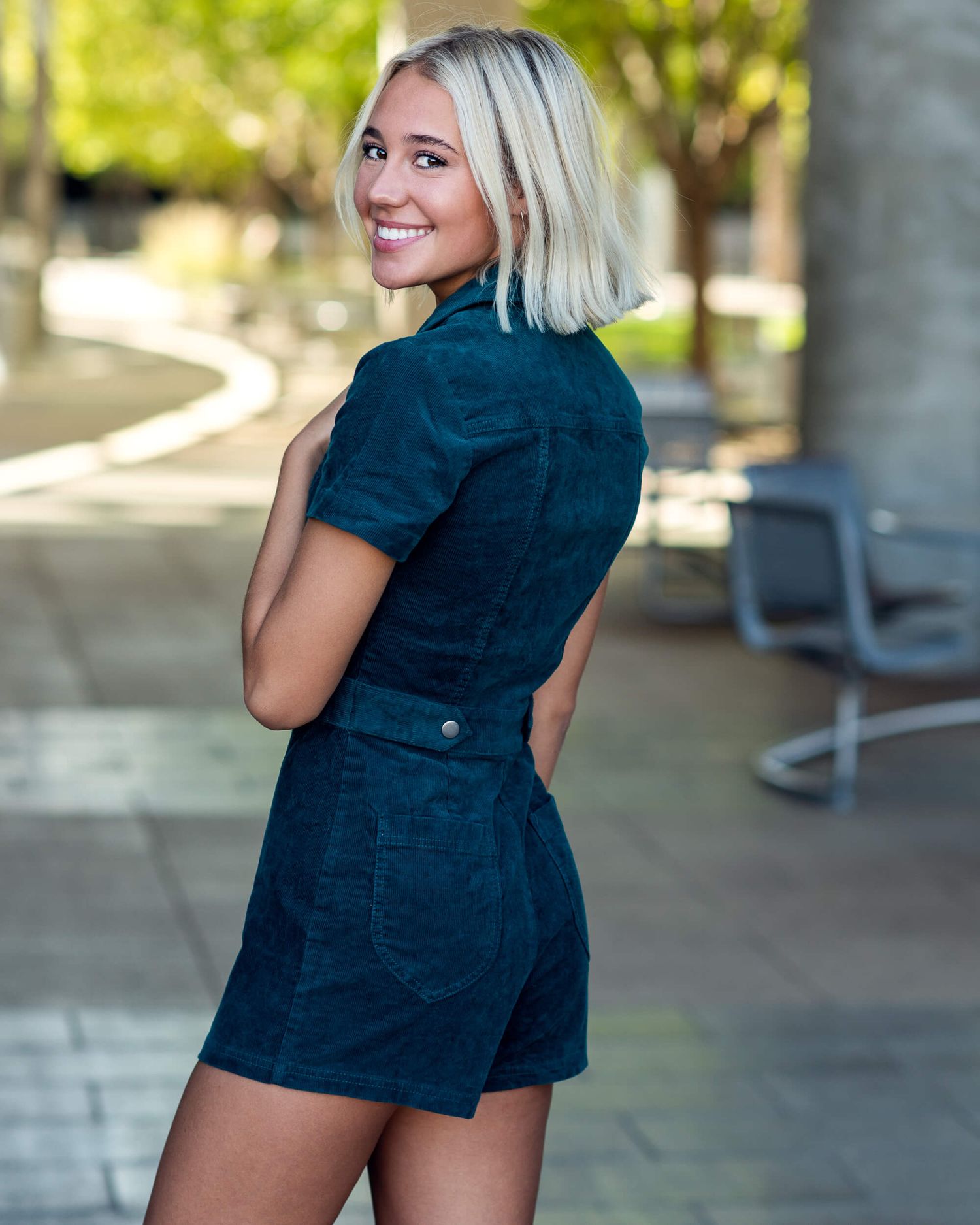 Blonde woman in a teal corduroy romper smiling over her shoulder outdoors.