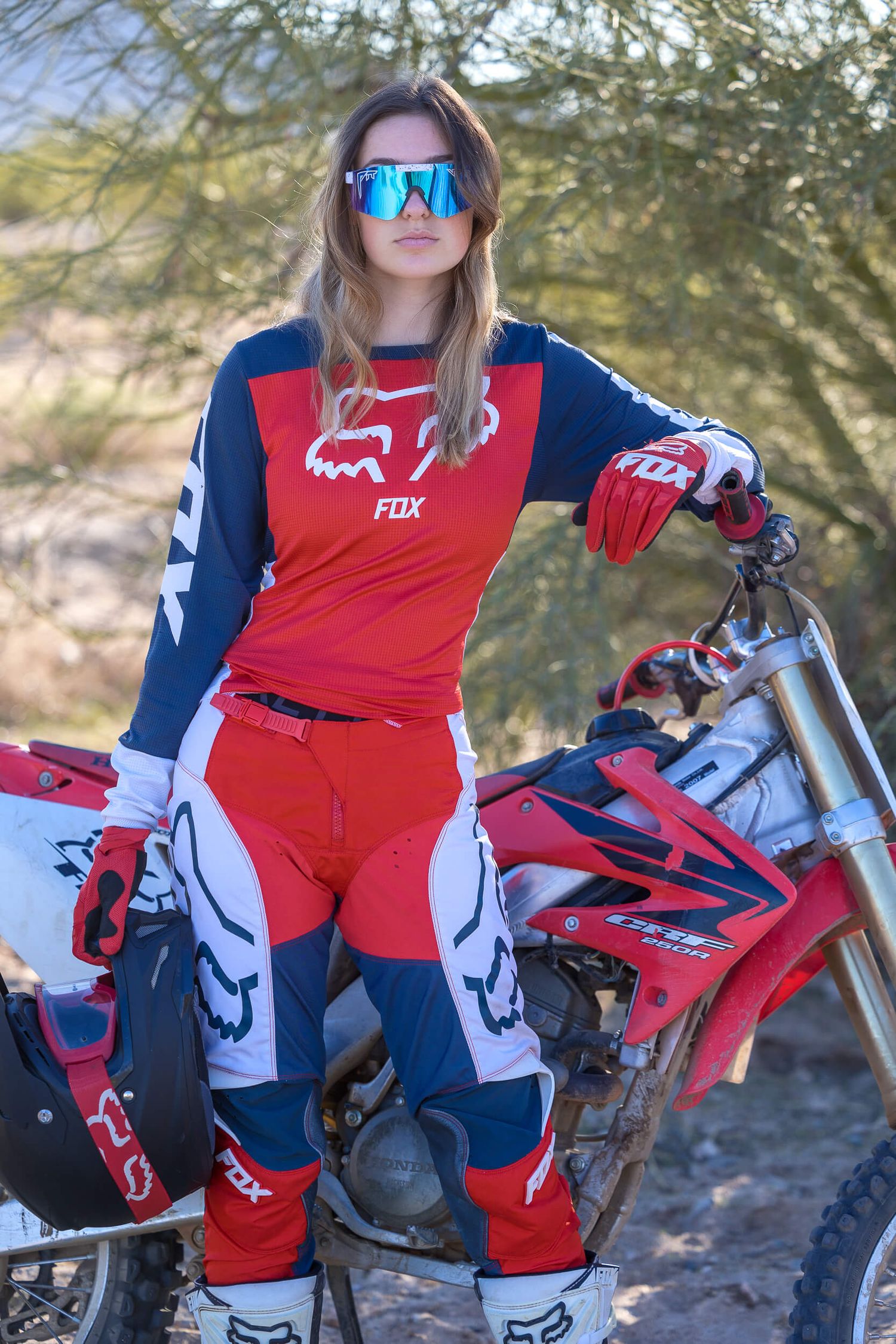 Woman in Fox Racing motocross gear posing with a dirt bike in a desert setting.