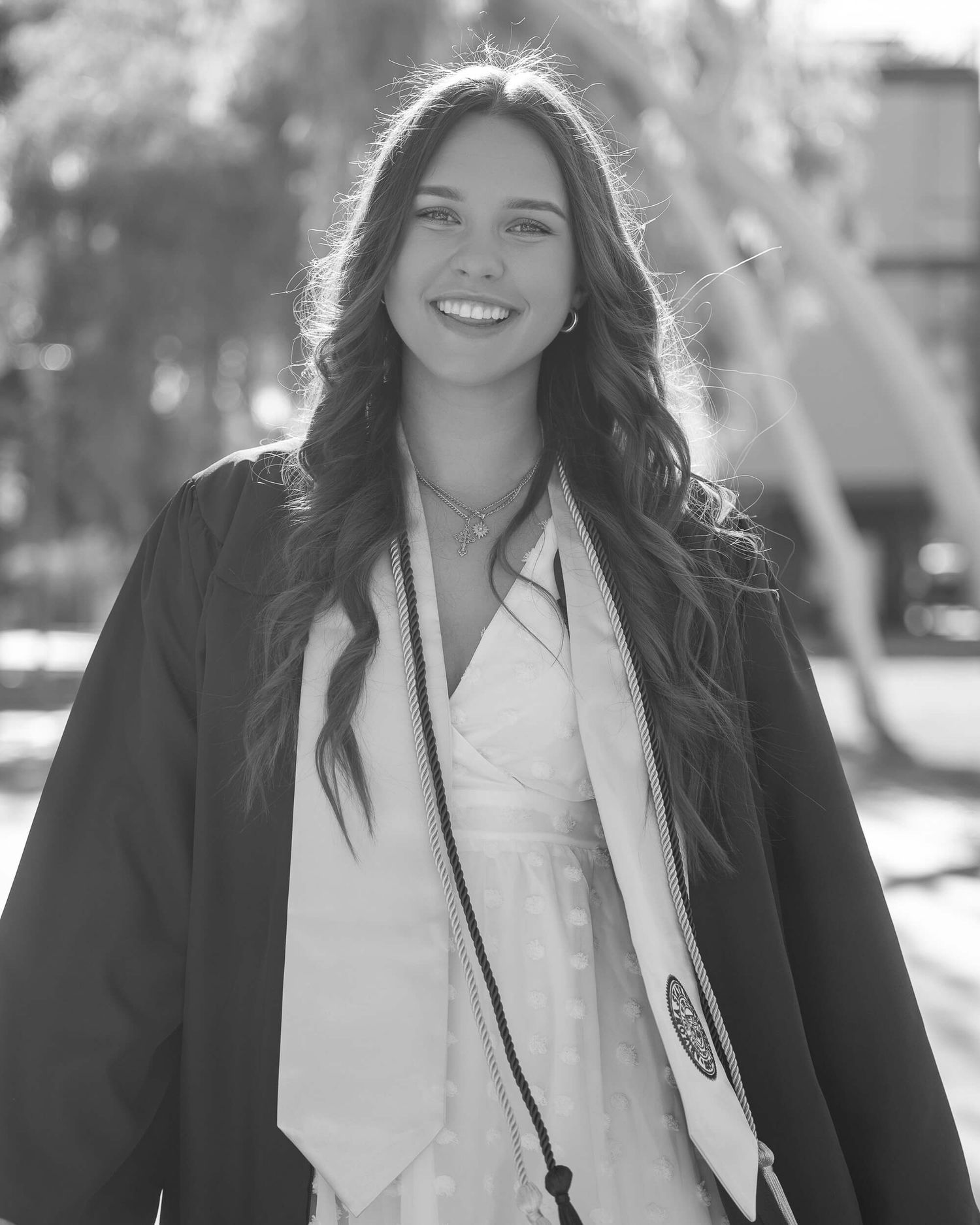 Black and white photo of smiling young woman in graduation gown and stole outdoors.