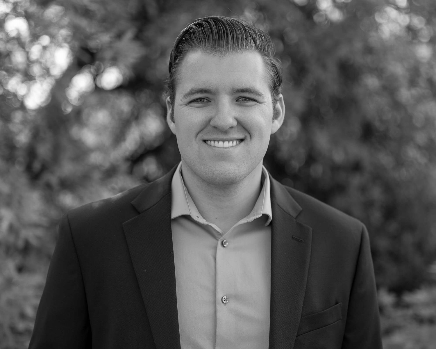 Professional black and white headshot of a smiling young man in a blazer outdoors.