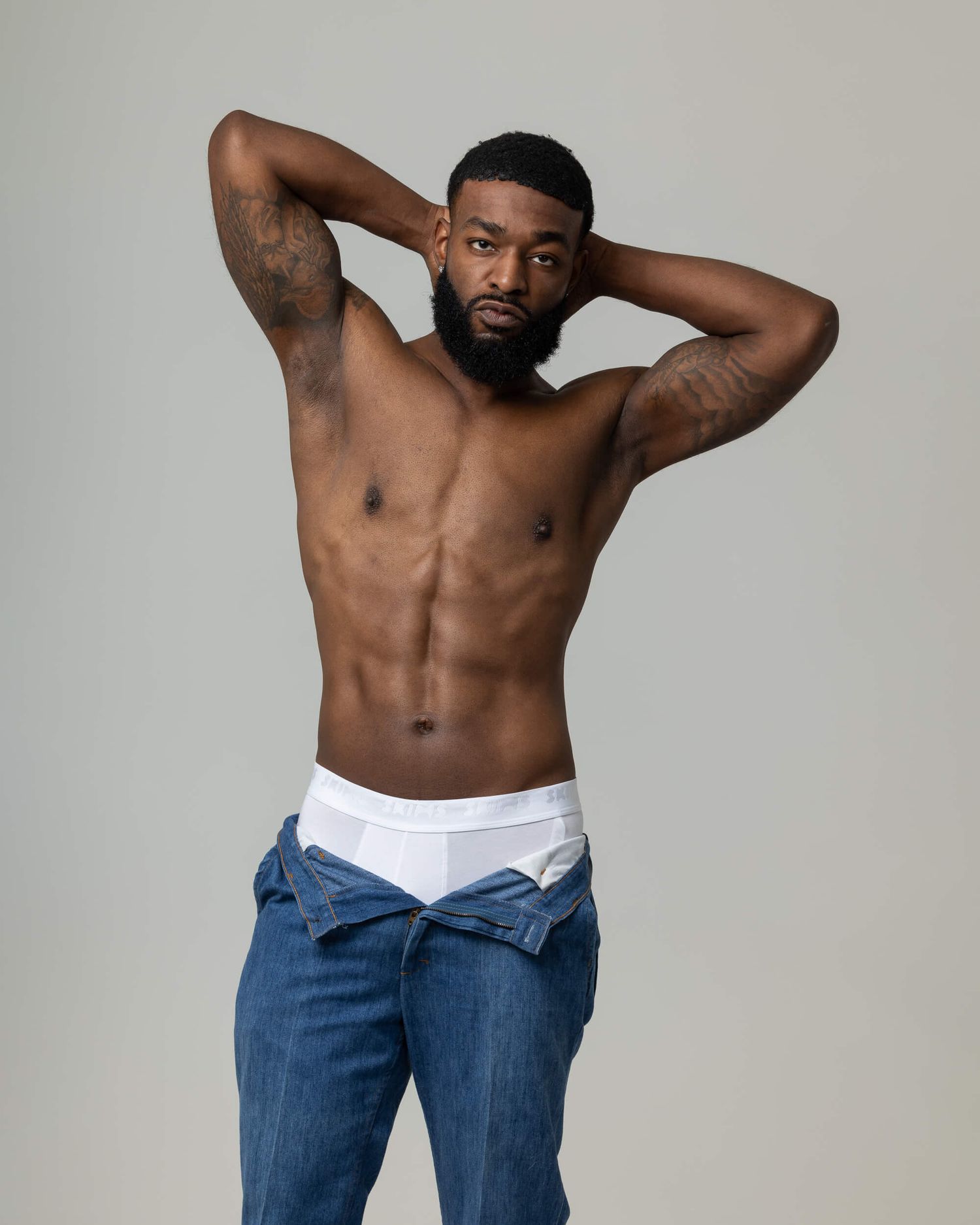 Shirtless muscular man with beard, tattoos, hands behind head, wearing white underwear and open jeans, studio photo.