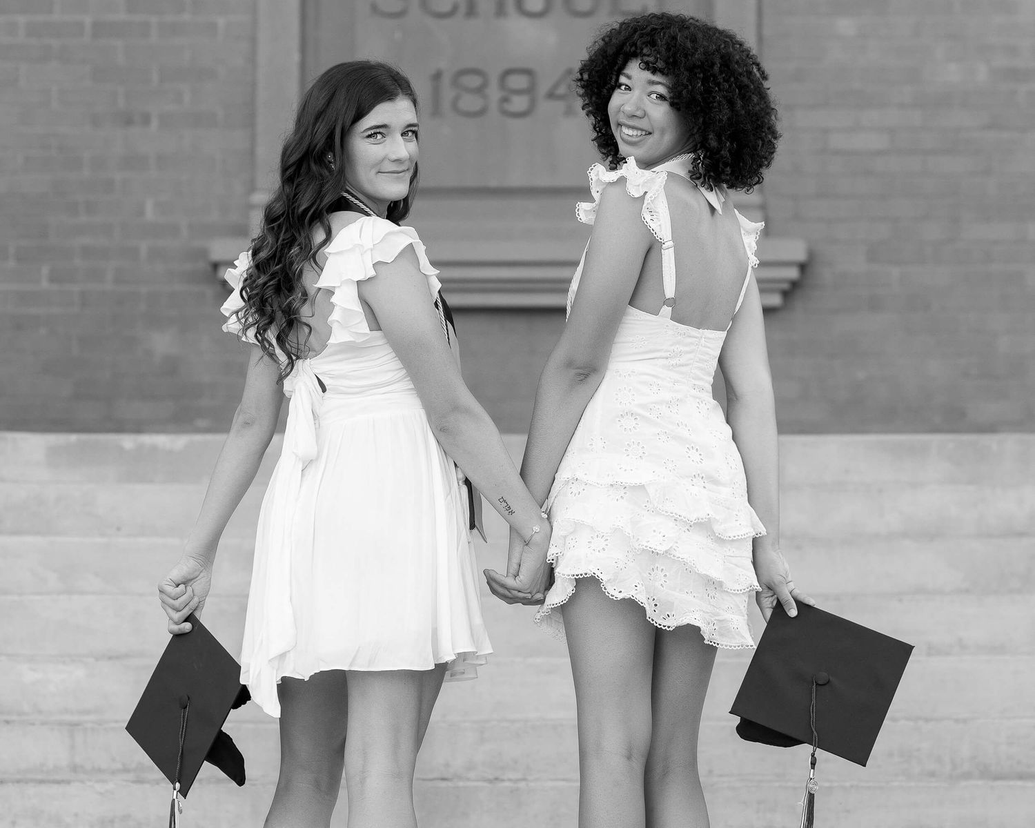 Black and white photo of two graduates in white dresses holding caps while facing away on campus steps.